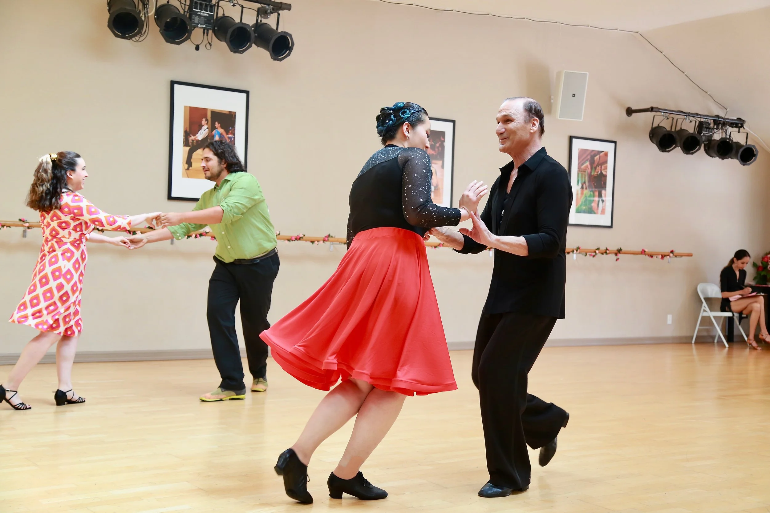 Couples dancing in a room decorated for a dance event. The woman in the foreground is wearing a red skirt and black top, dancing with a man in a black shirt. In the background, another couple is holding hands and dancing, with the woman in a colorful patterned dress and the man in a light green shirt. The room has framed pictures on the wall, stage lights hanging from the ceiling, and a woman sitting on a chair.