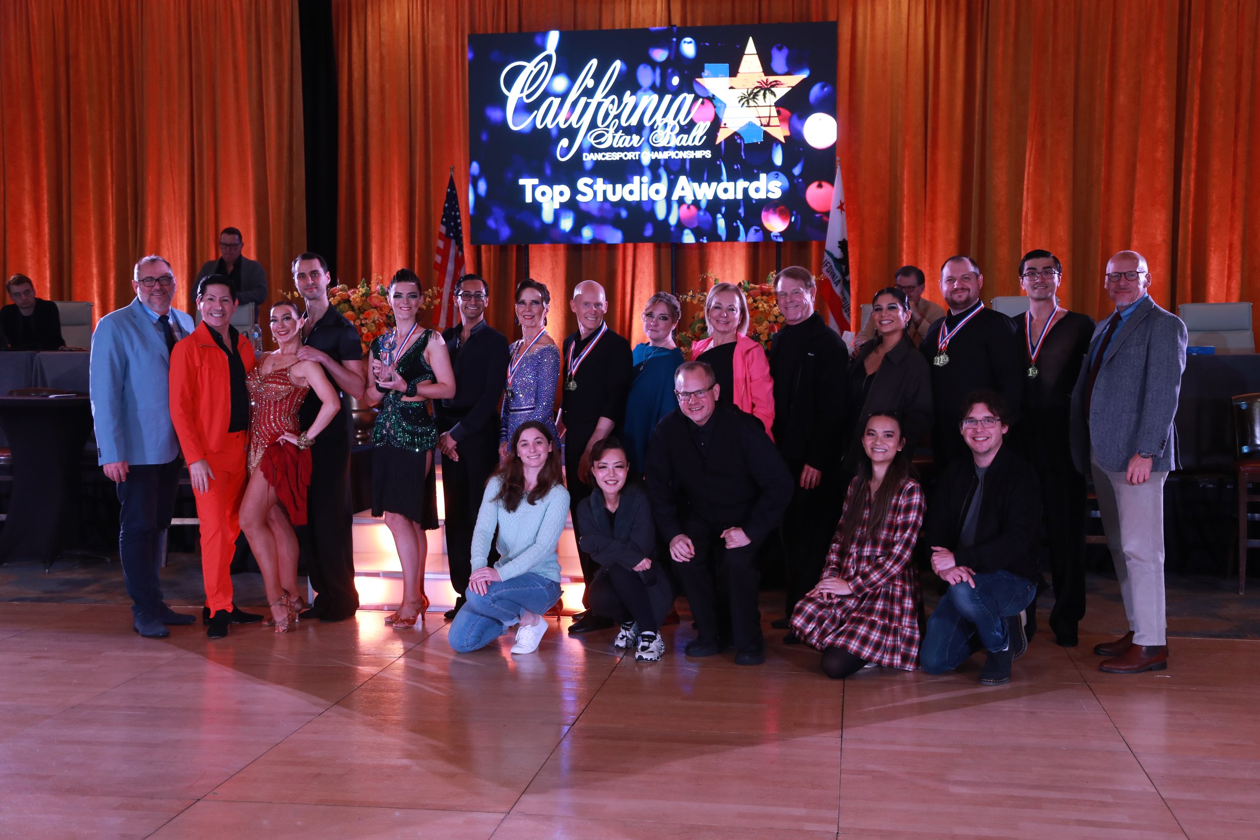 Group of dancers and their instructors smiling for a photo at the California Star Ball dance competition awards ceremony, with a large digital screen displaying the event name and awards in the background.