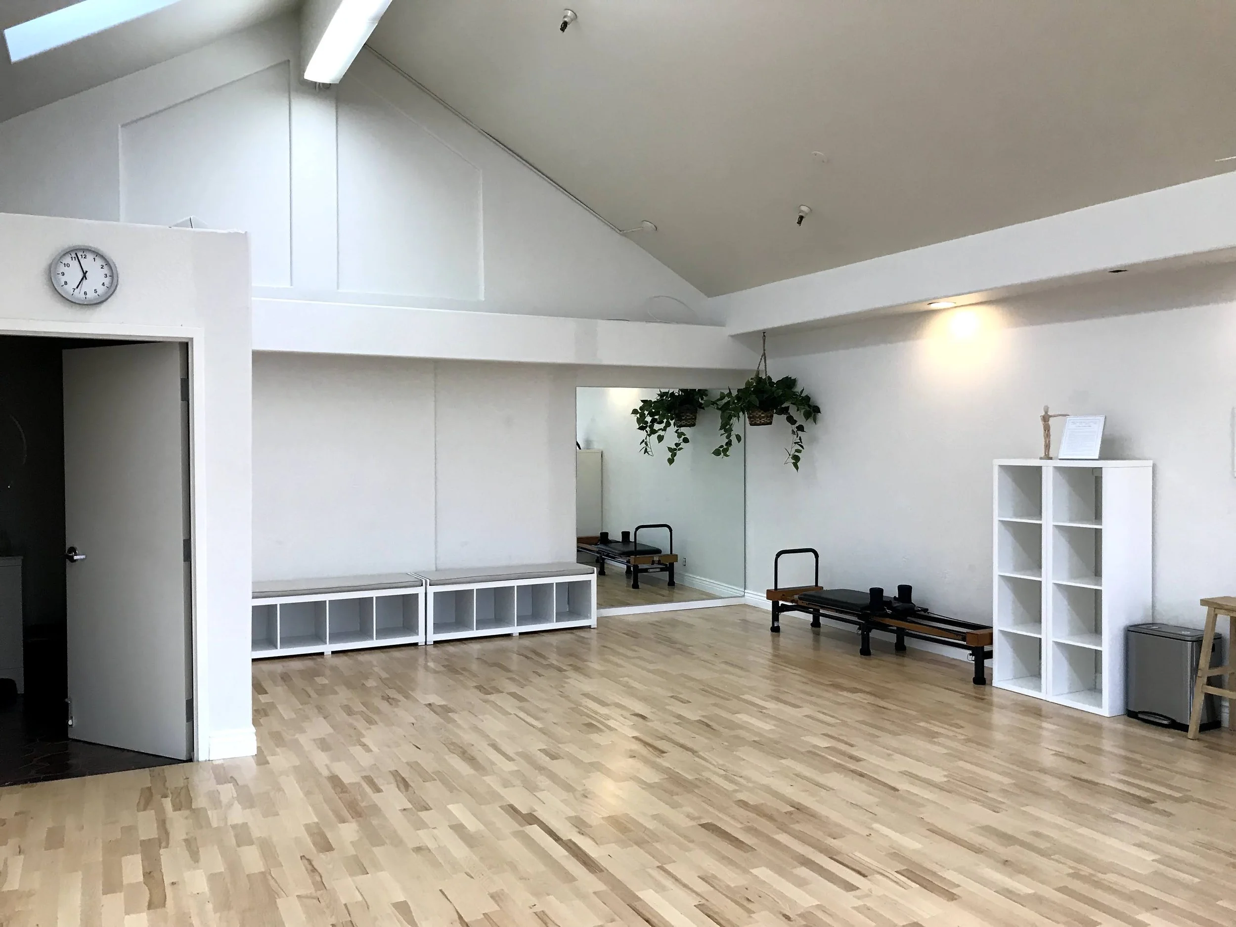 A spacious room with light-colored hardwood flooring, white walls, and high ceilings. There are mirrors on the wall, potted plants hanging from the ceiling, and furniture including benches and shelves. A clock is mounted on the wall near a doorway.