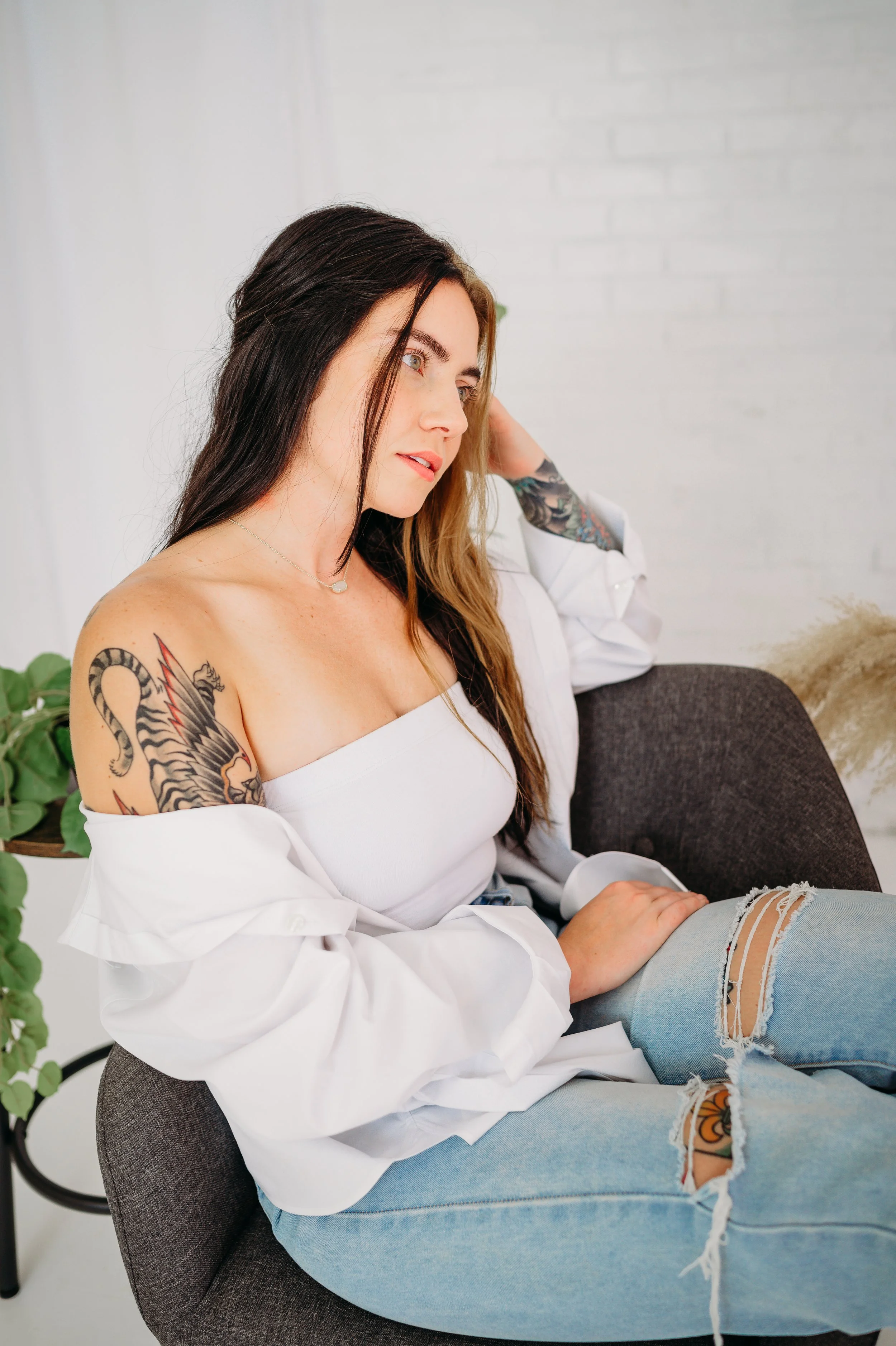 A young woman sitting on a gray chair, wearing a white off-the-shoulder top and ripped jeans, with tattoos on her arm, in a bright room with white walls and green plants.