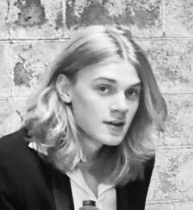 Portrait of a young woman with wavy blonde hair, wearing a dark blazer and a light-colored top, standing in front of a textured brick wall.