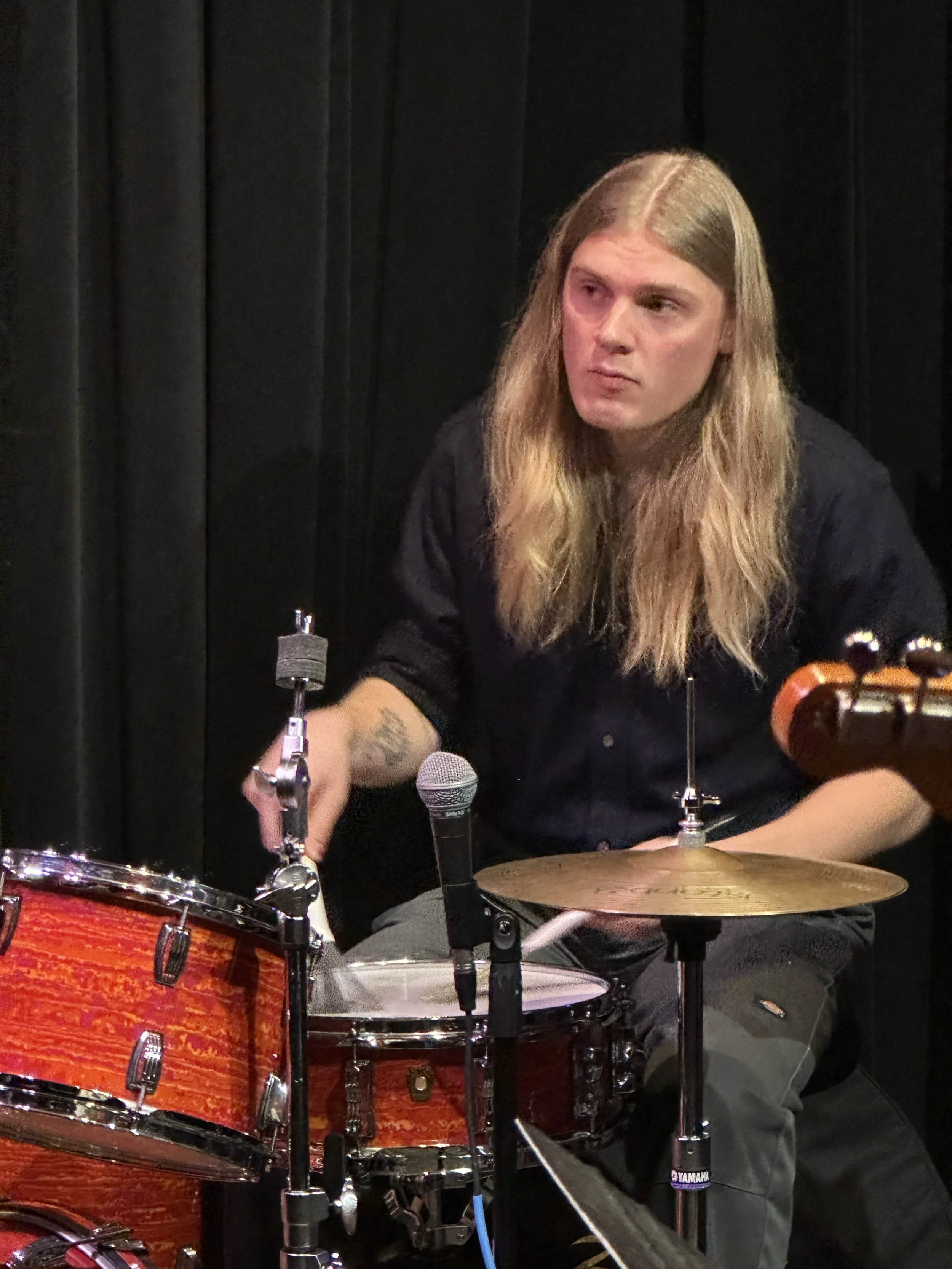 A man with long blond hair playing a drum set, with a black curtain background, wearing a black shirt and sitting behind drums with drumsticks in hand.