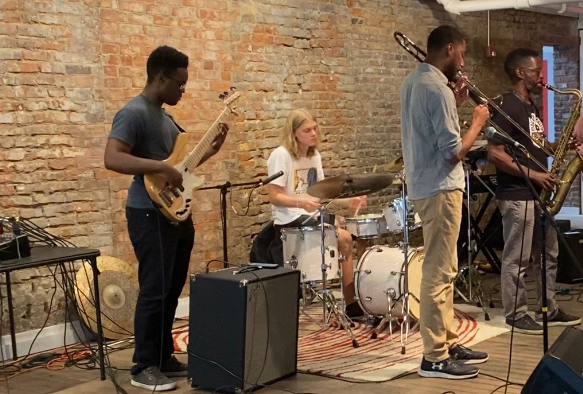A band of four musicians performing live in an indoor venue with a brick wall background. The band includes a guitarist, a drummer, and two saxophonists.