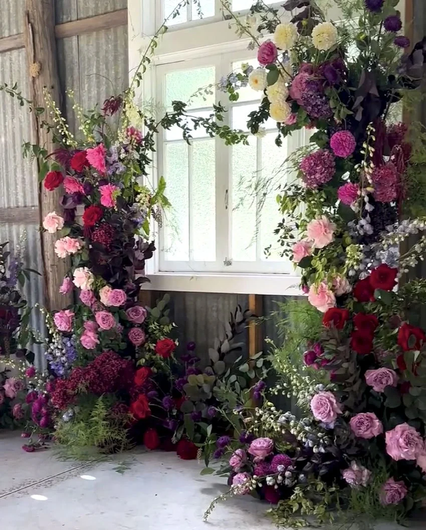 A floral arch made of pink, purple, red, and white flowers and green foliage, set up in front of a window with frosted glass panes, inside a rustic space with corrugated metal walls and a wooden backdrop.
