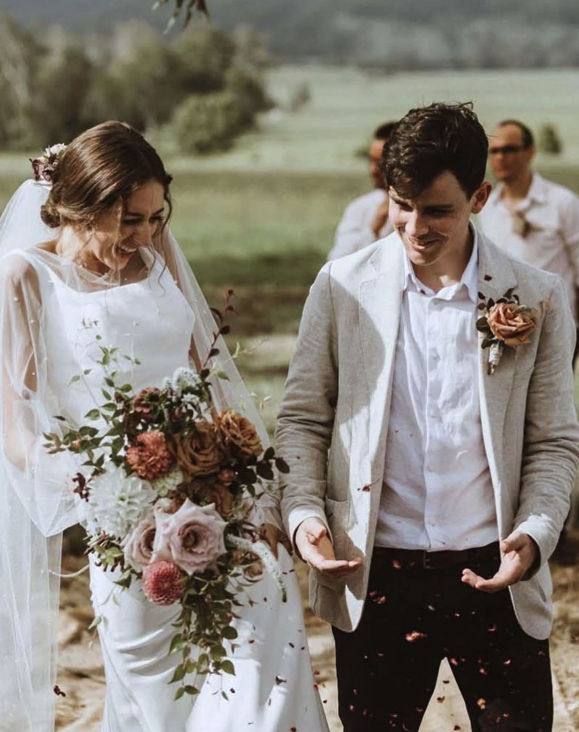 A bride and groom walking outdoors during their wedding, holding hands and smiling with wedding guests in the background.