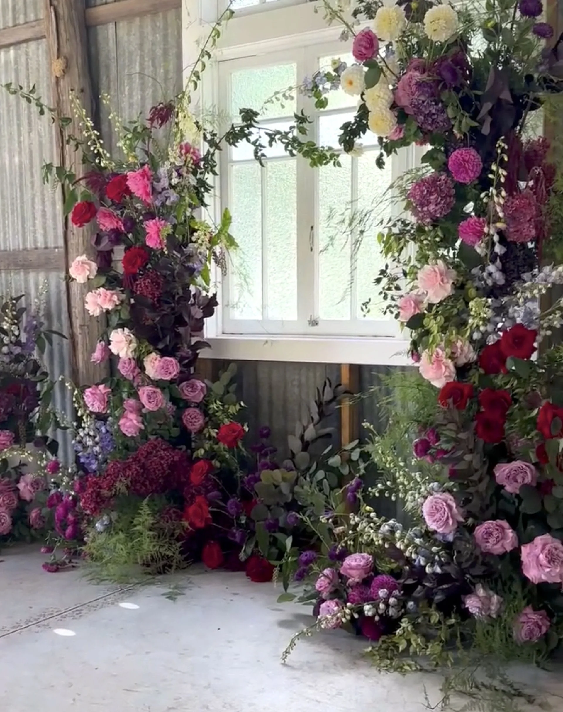 A floral decoration with various pink, purple, white, and red flowers on a circular floral arch inside a rustic barn or shed with metal siding and a window.