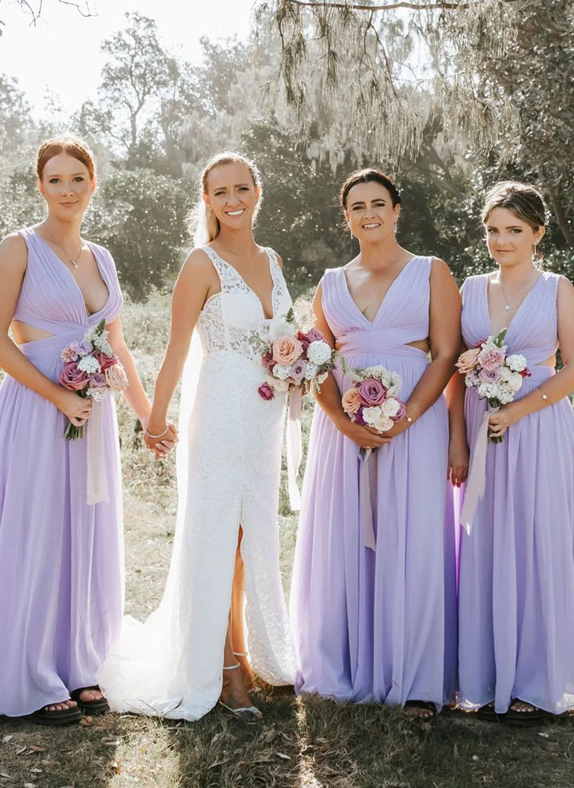 A bride in a white wedding dress holding hands with bridesmaids dressed in lavender gowns, all holding bouquets of pink and white roses outdoors.