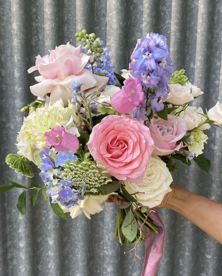 A hand holding a bouquet of pastel-colored flowers including pink, white, blue, and purple blooms, with green leaves and a pink ribbon, against a gray curtain background.