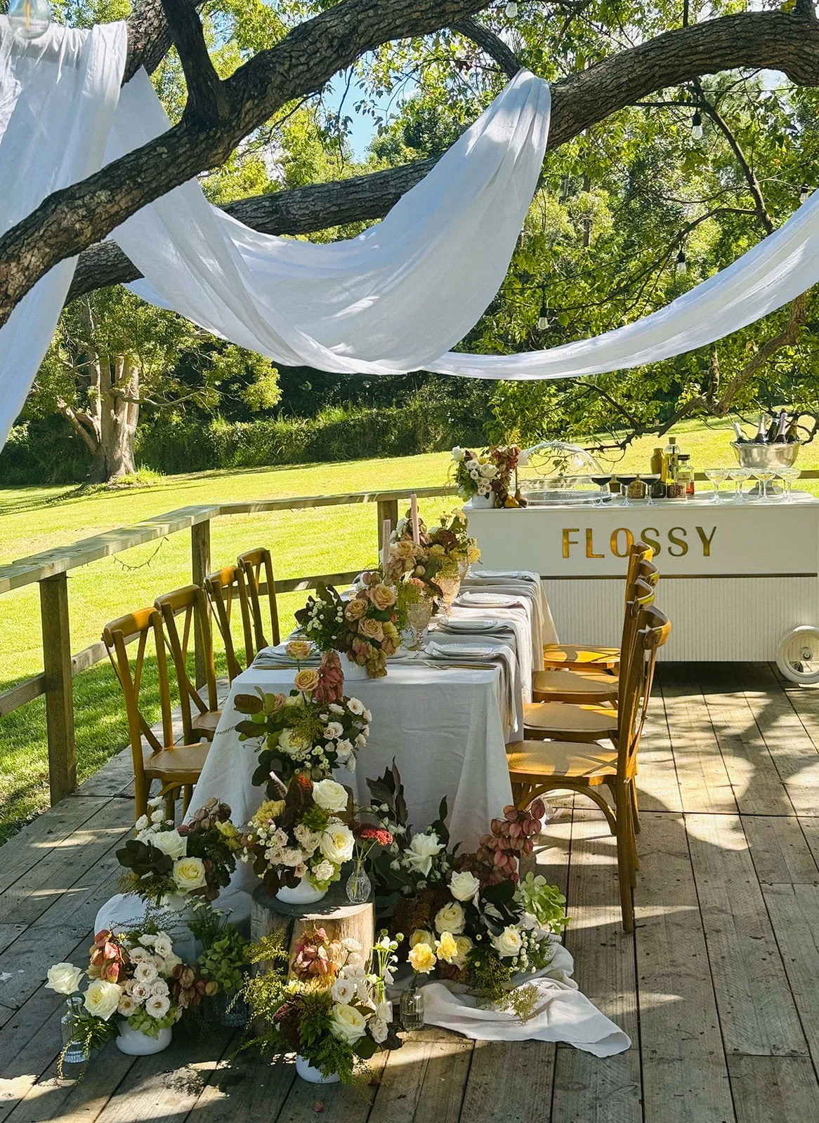 Outdoor wedding or event setting with a long table decorated with flowers, a white tablecloth, and chairs. Behind the table, there's a white cart with gold letters spelling "FLOSSY" and various bottles. A tree with white drapes and string lights prov