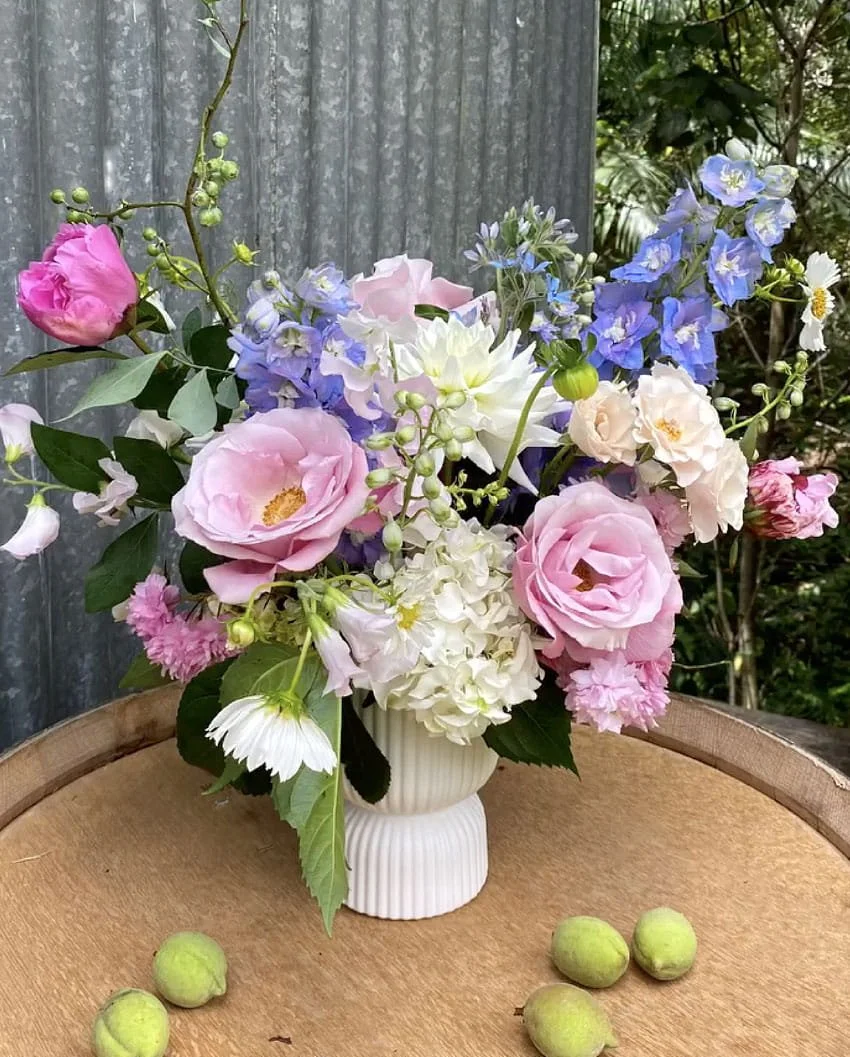 A bouquet of pink, white, and blue flowers in a white vase on a wooden surface, with small green fruits scattered around.