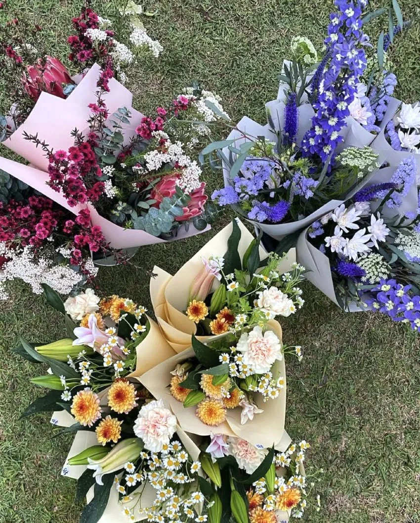 Multiple colorful flower bouquets on grass, including pink, white, purple, yellow, and orange blossoms wrapped in paper.