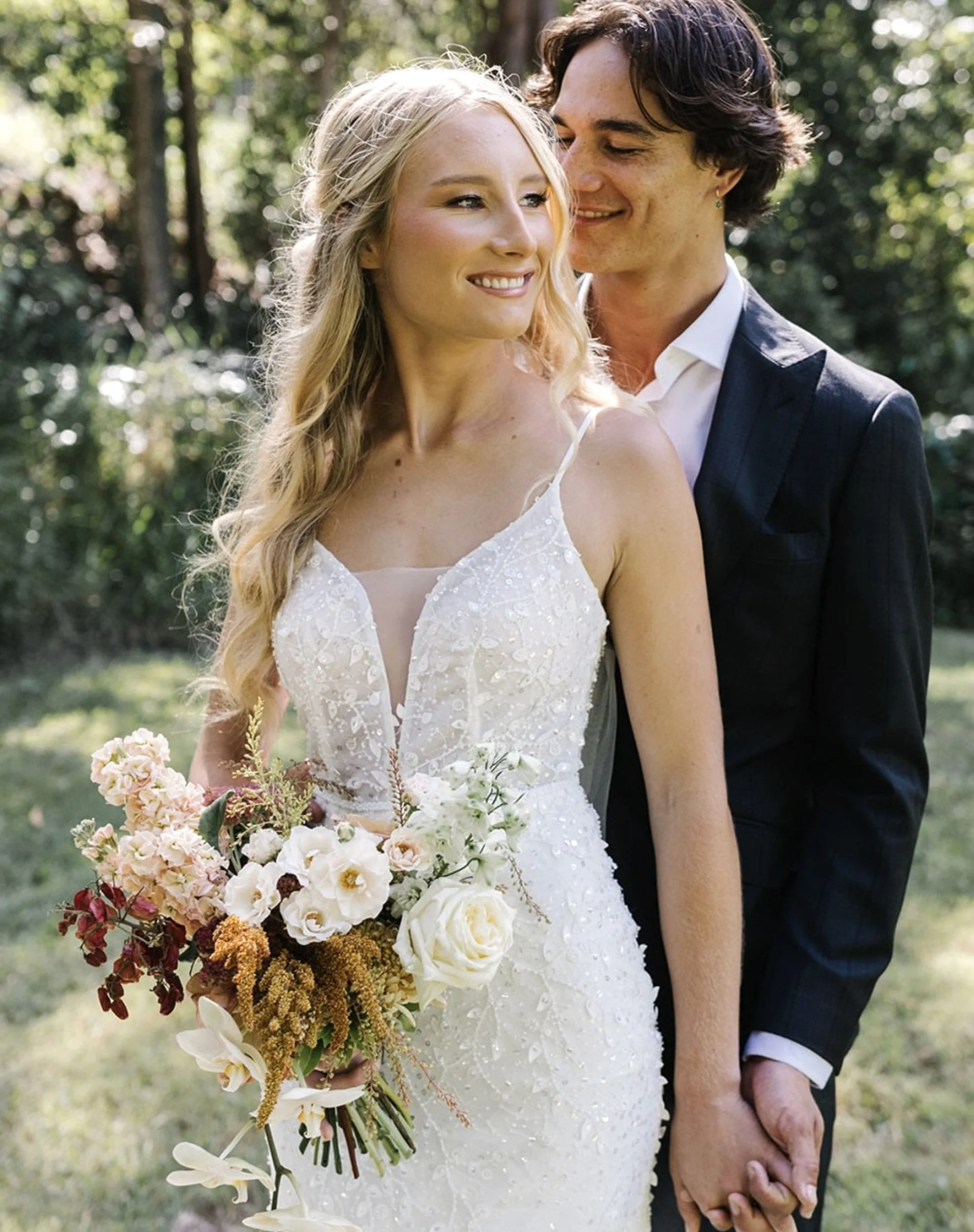 A bride and groom holding hands outdoors during their wedding. The bride has long blonde hair, wearing a white wedding dress and holding a large bouquet of flowers. The groom has dark hair, wearing a dark suit, white shirt, and is smiling at the brid