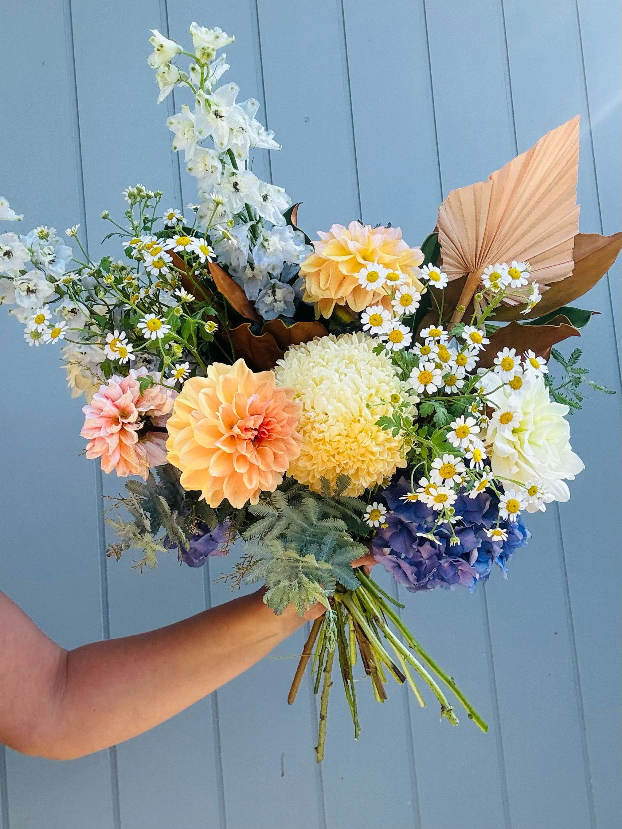 A person holding a bouquet of mixed flowers, including white, yellow, pink, and purple blooms, against a blue wooden background.