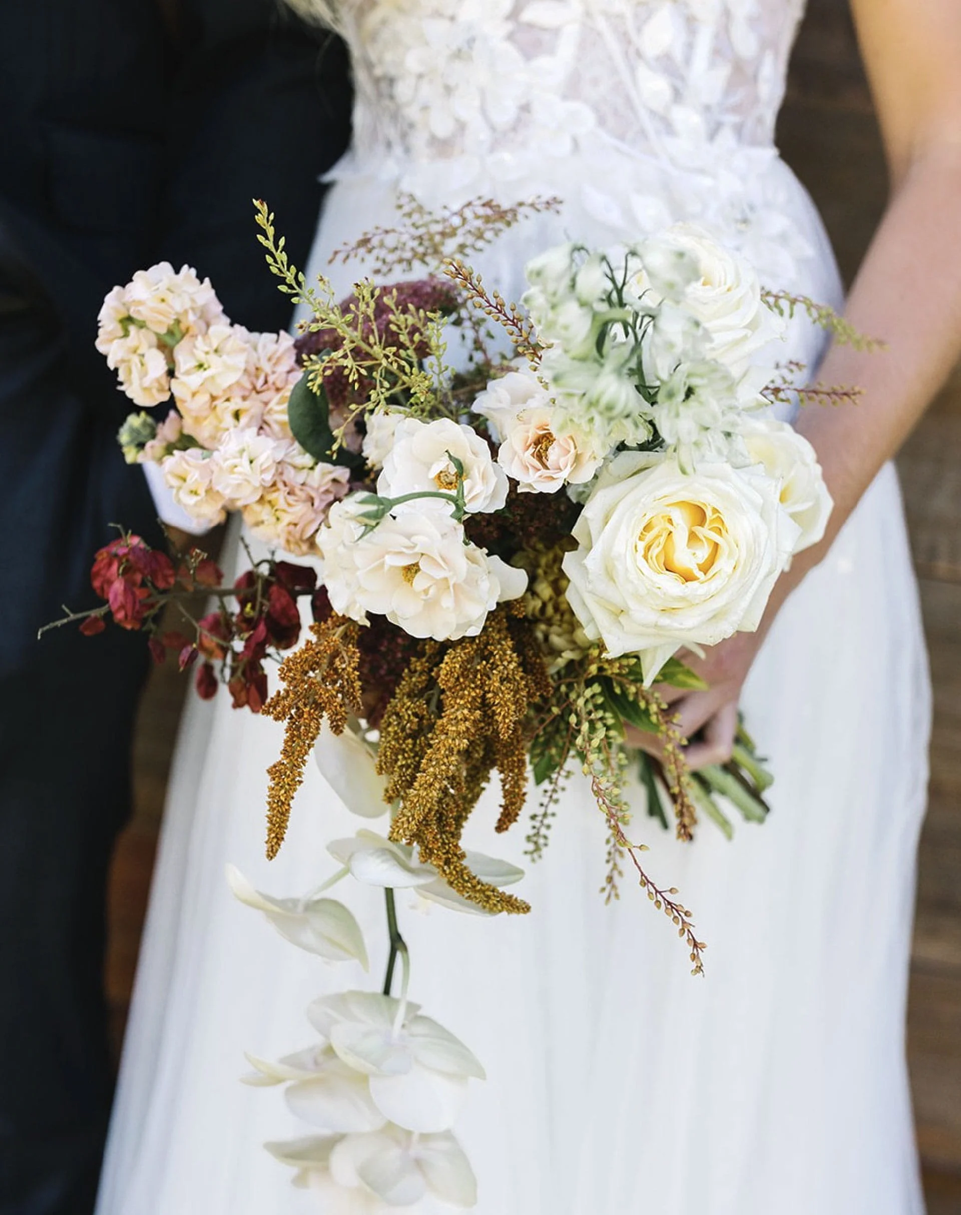 Close-up of a bride holding a bouquet of white, peach, and burgundy flowers with greenery, against a white dress. The background is blurred.
