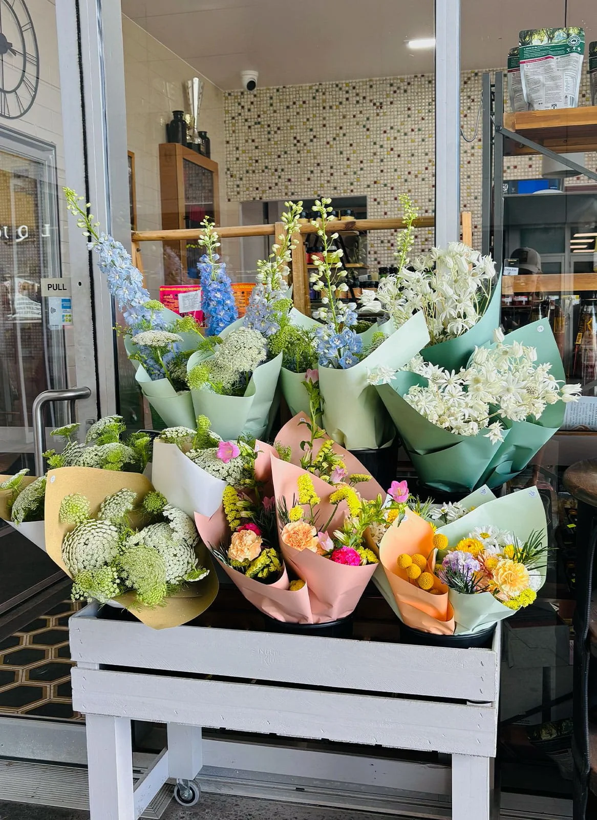 A display of colorful flower bouquets arranged in a shopping cart outside a store, with a mosaic wall and shelving in the background.