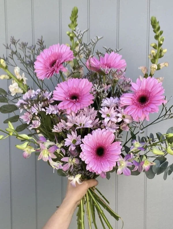 Hand holding a bouquet of pink and purple flowers with green foliage in front of a light gray background.