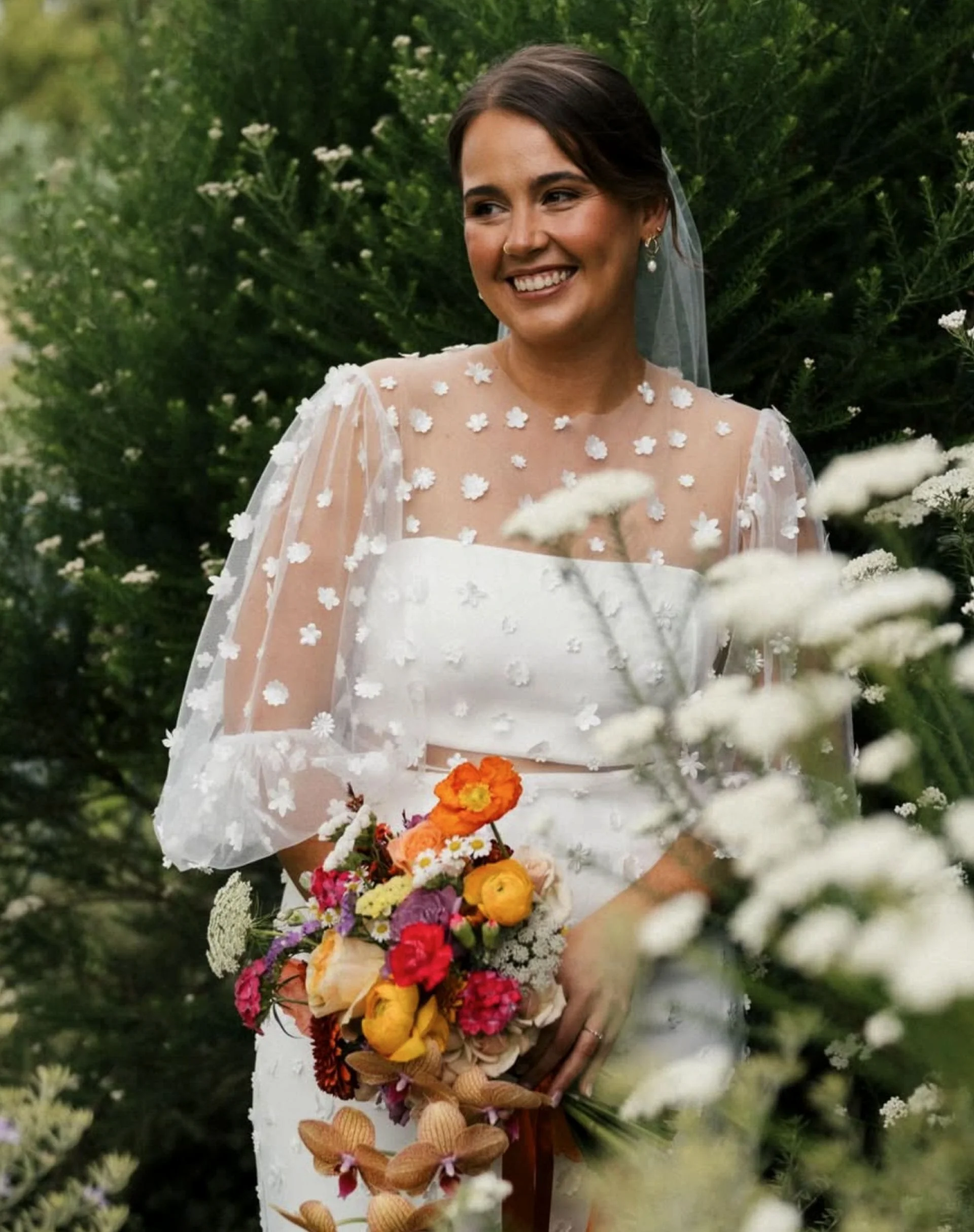 A smiling woman in a wedding dress holding a colorful bouquet of flowers, standing outdoors among green trees and white flowers.