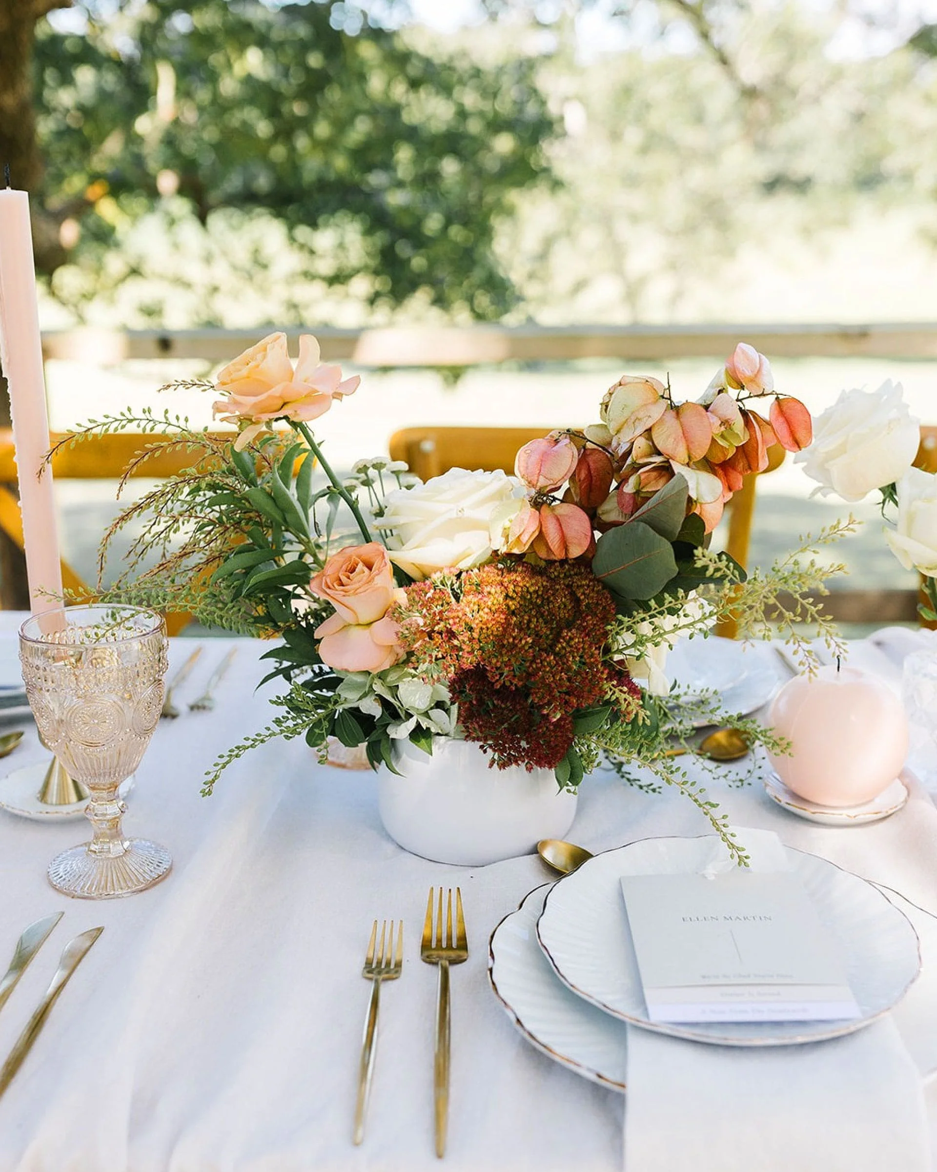 A table decorated with a floral centerpiece, gold utensils, and elegant glassware, set for a formal event outdoors with blurred trees in the background.