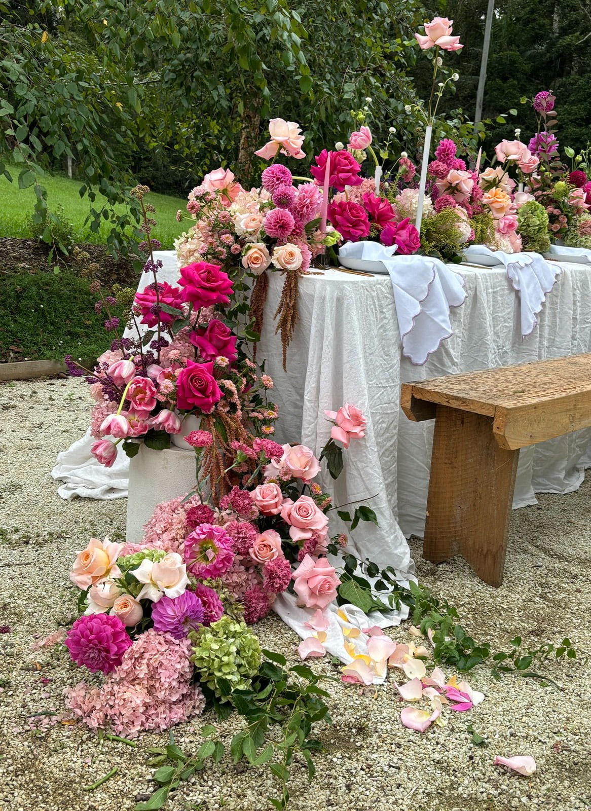 A table decorated with a white cloth and surrounded by pink and peach roses, hydrangeas, and other flowers for a wedding or special event, with petals scattered on the ground.