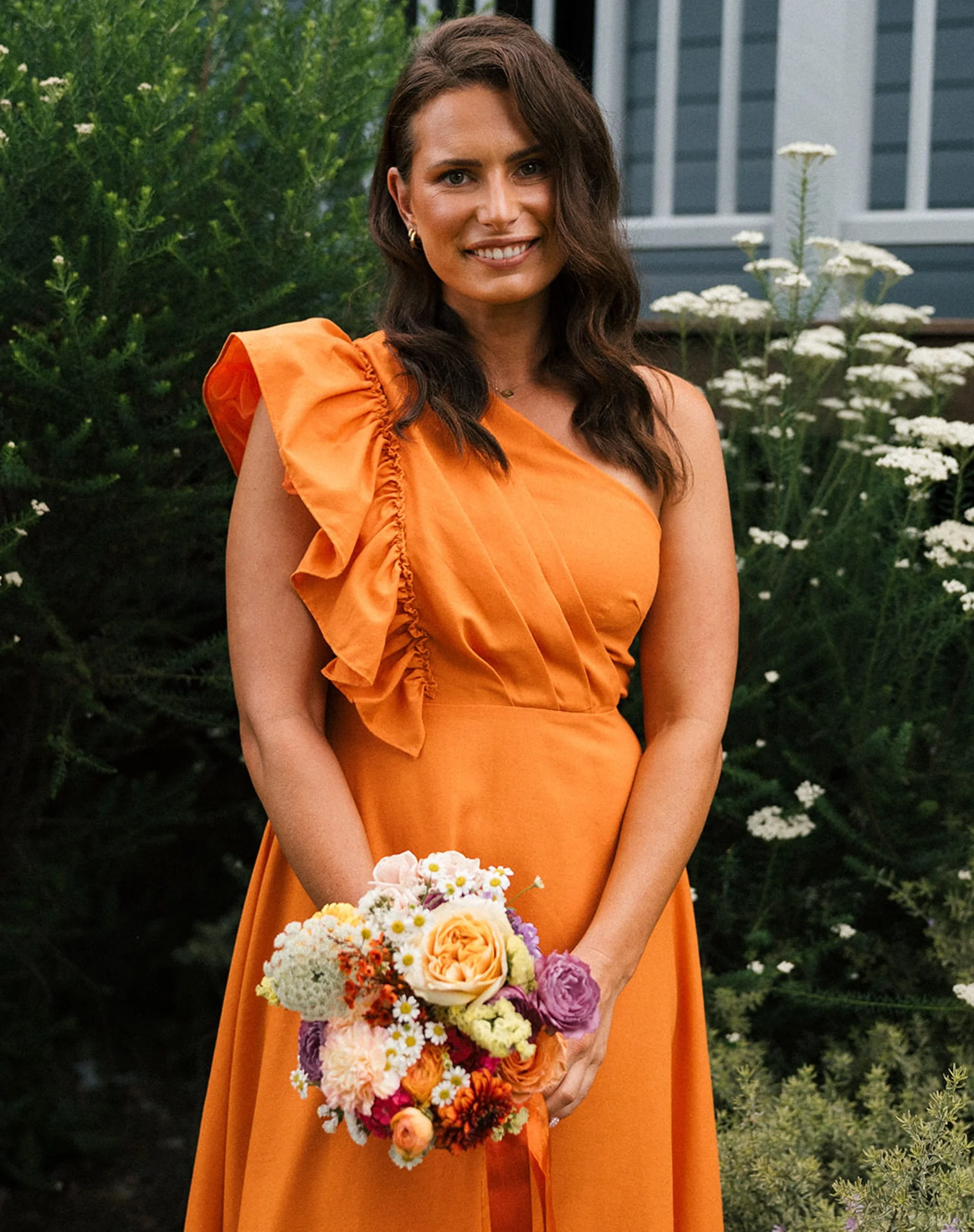 A woman with long brown hair wearing an orange dress and holding a colorful bouquet of flowers standing outdoors in a garden with greenery and white flowers.