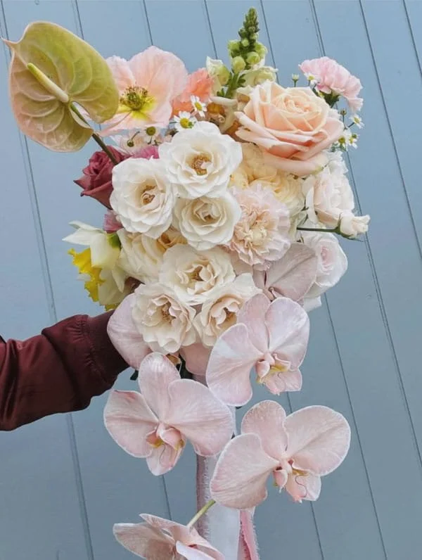 A person holding a large bouquet of pastel-colored flowers, including roses, orchids, and other blooms, against a blue wooden background.