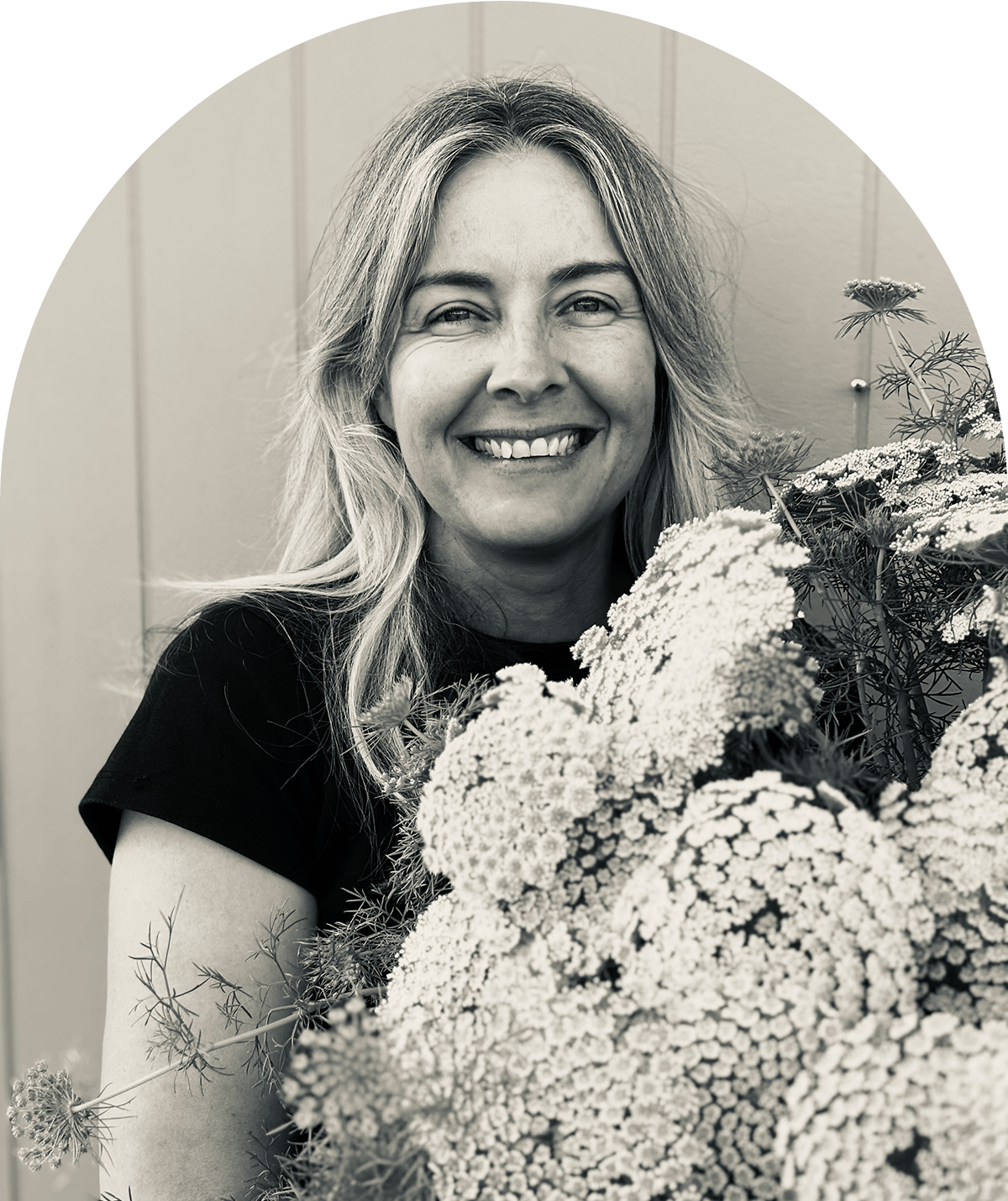 A smiling woman with wavy blonde hair holding a large bouquet of flowers, with a light-colored wall in the background. Renee Gillbank.