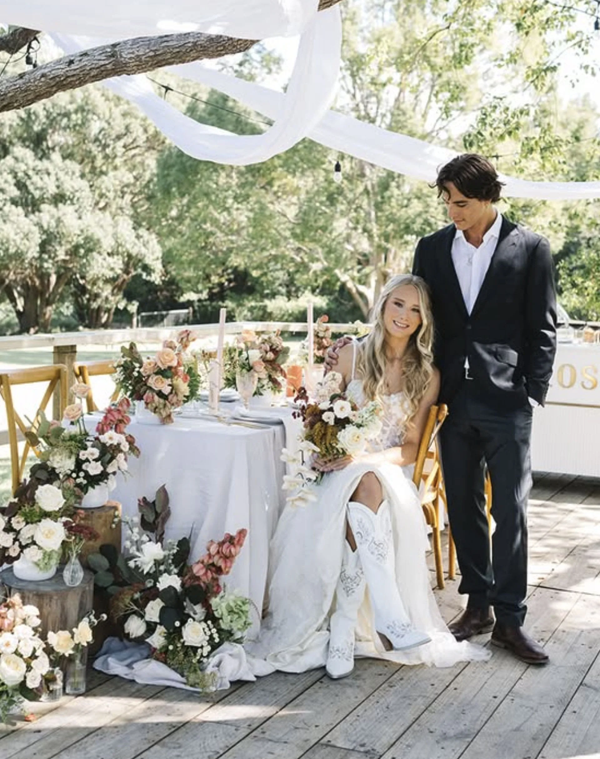 A wedding scene outdoors with a bride and groom, floral decorations on a table, and decorative white fabric hanging from trees.