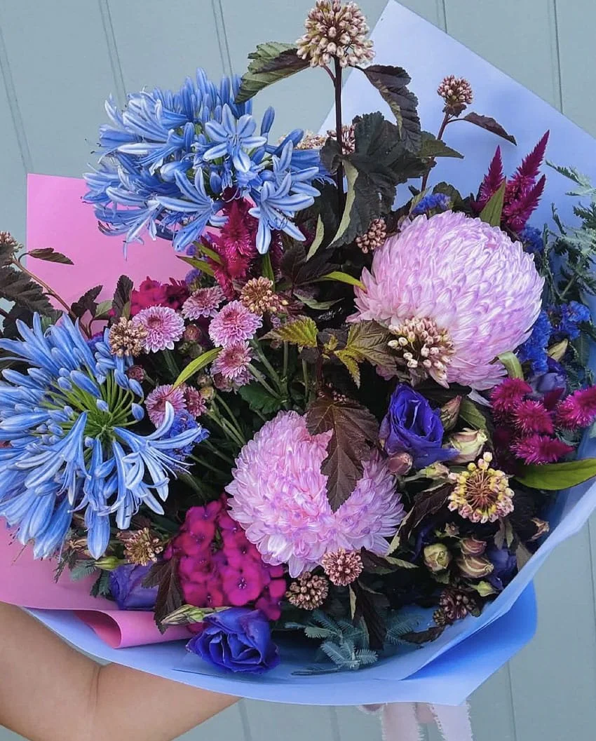 A colorful bouquet of mixed flowers including pink dahlias, blue agapanthus, purple lisianthus, pink chrysanthemums, and other assorted blooms wrapped in pink and blue paper.
