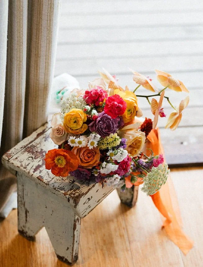 Colorful bouquet of mixed flowers placed on a rustic, white, distressed wooden stool near window with curtains.