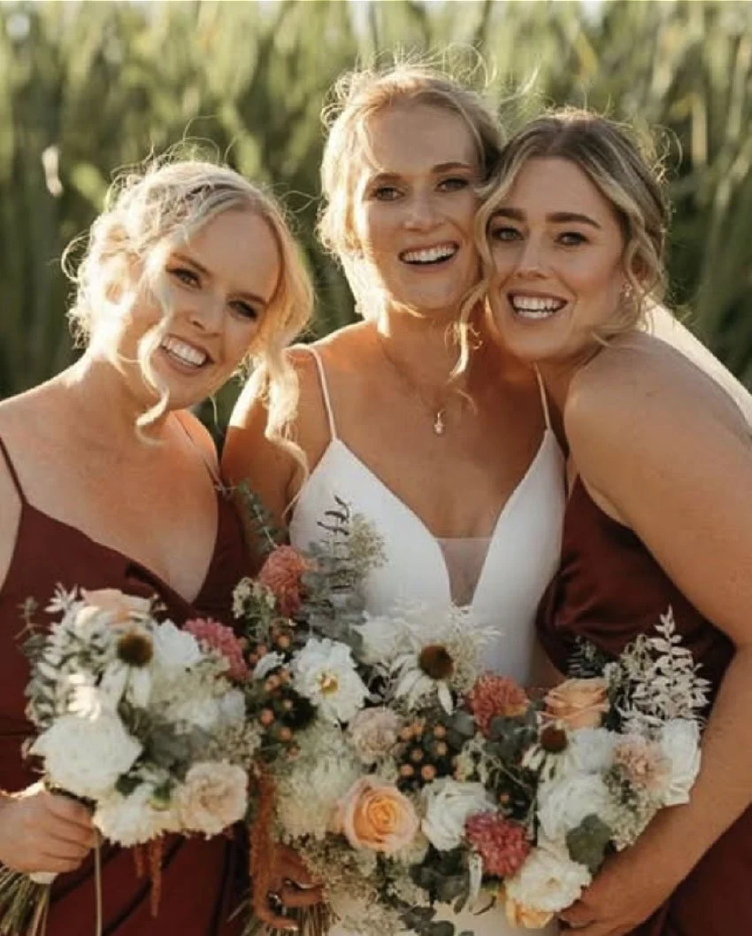 Three women smiling and posing together outdoors during a wedding, holding large bouquets of flowers, with a background of greenery.