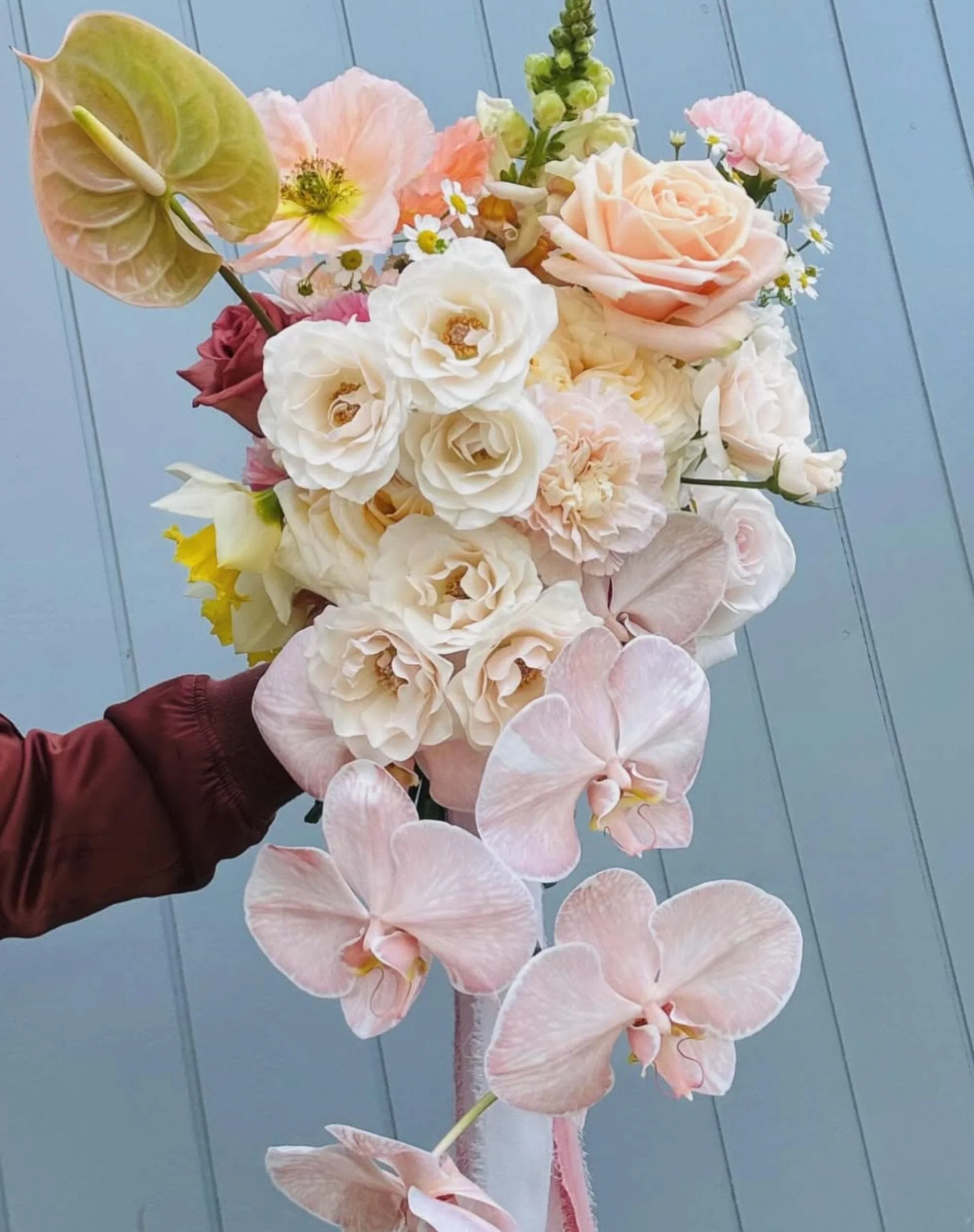 A person wearing a maroon jacket holding a large bouquet of mixed flowers including roses, orchids, and some greenery against a blue wooden background.