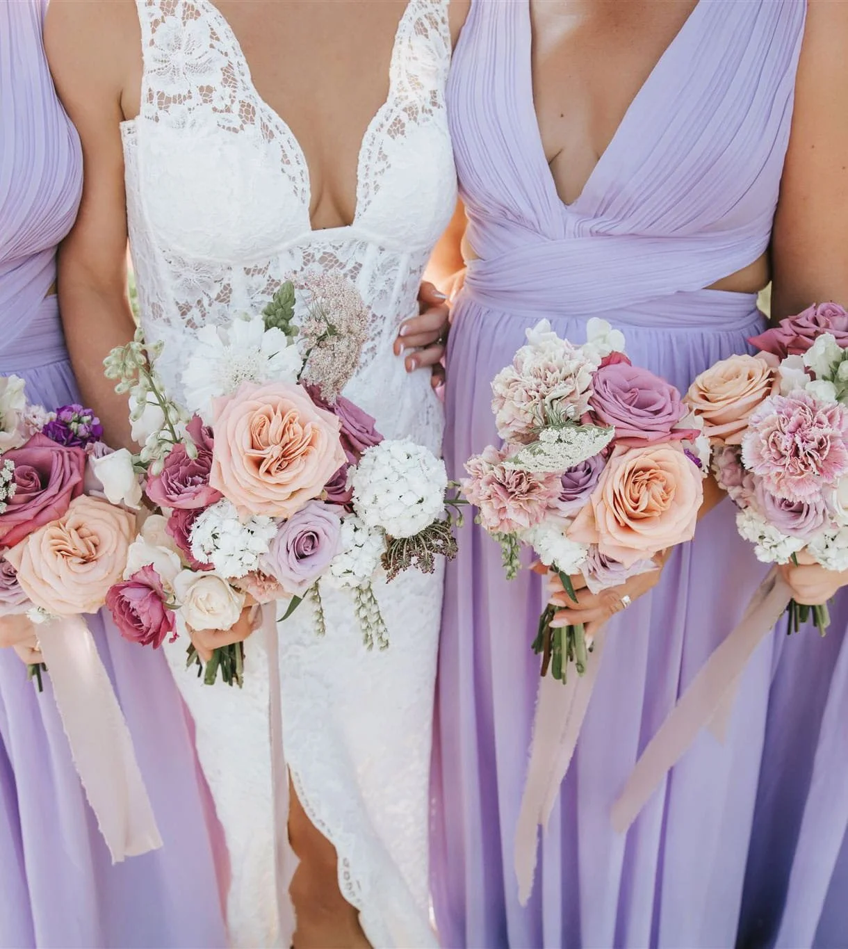 Close-up of a bride and bridesmaids holding bouquets of pink, purple, and white flowers, with the bride in a white lace dress and bridesmaids in lavender dresses.