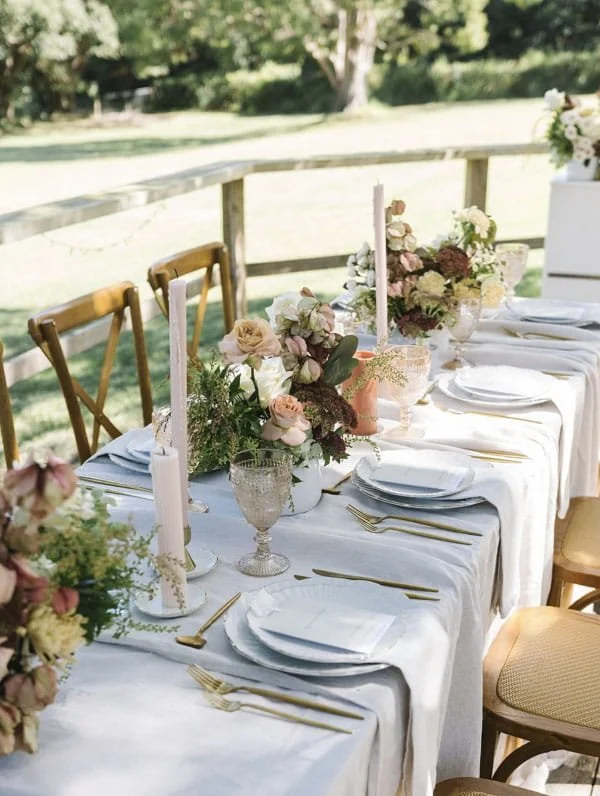 Outdoor wedding reception table with white tablecloths, floral centerpieces, white candles, gold flatware, and glassware, set on a porch overlooking a grassy yard and trees.