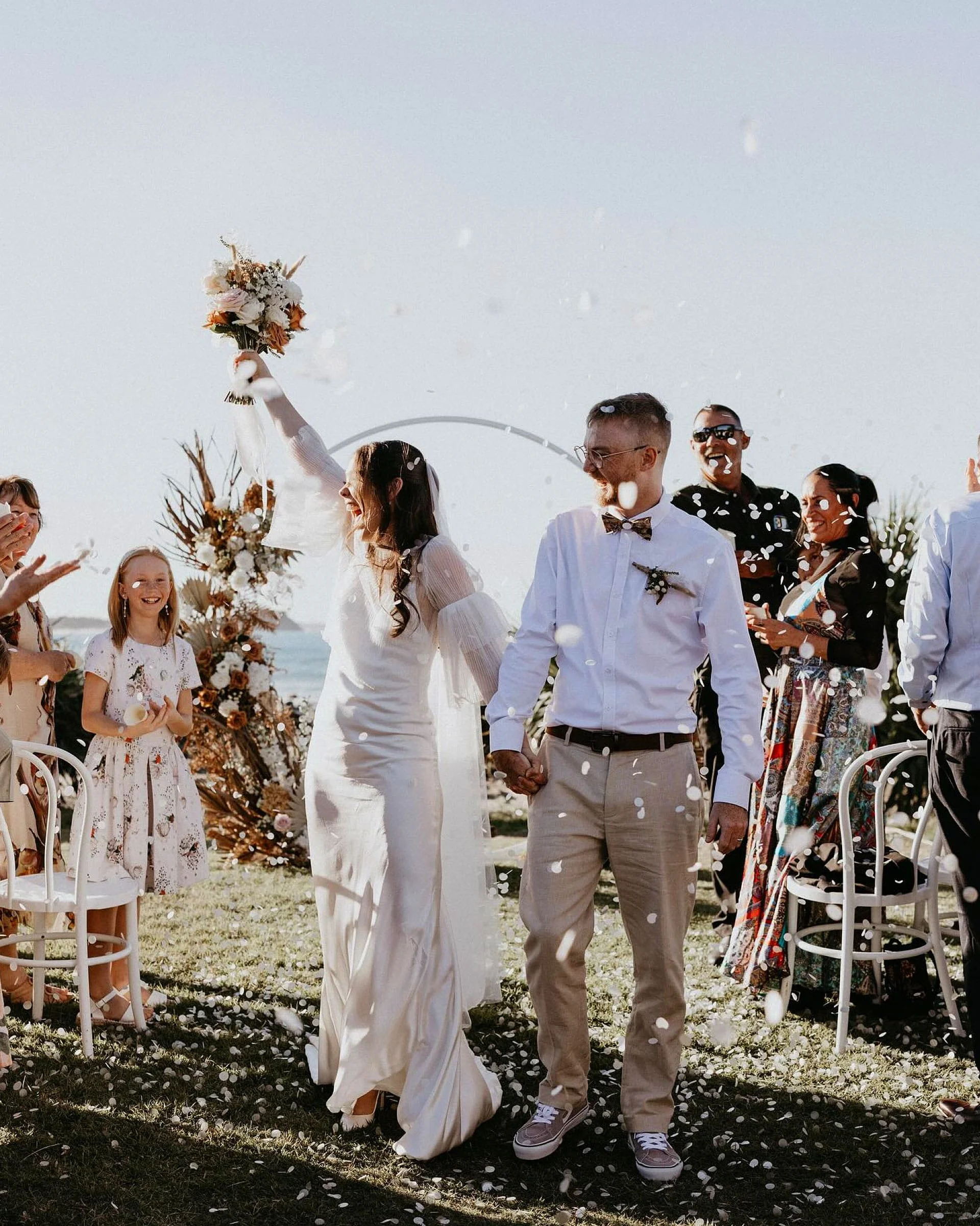 A wedding ceremony outdoors with a bride and groom holding hands, surrounded by friends and family, with floating confetti and floral decorations.