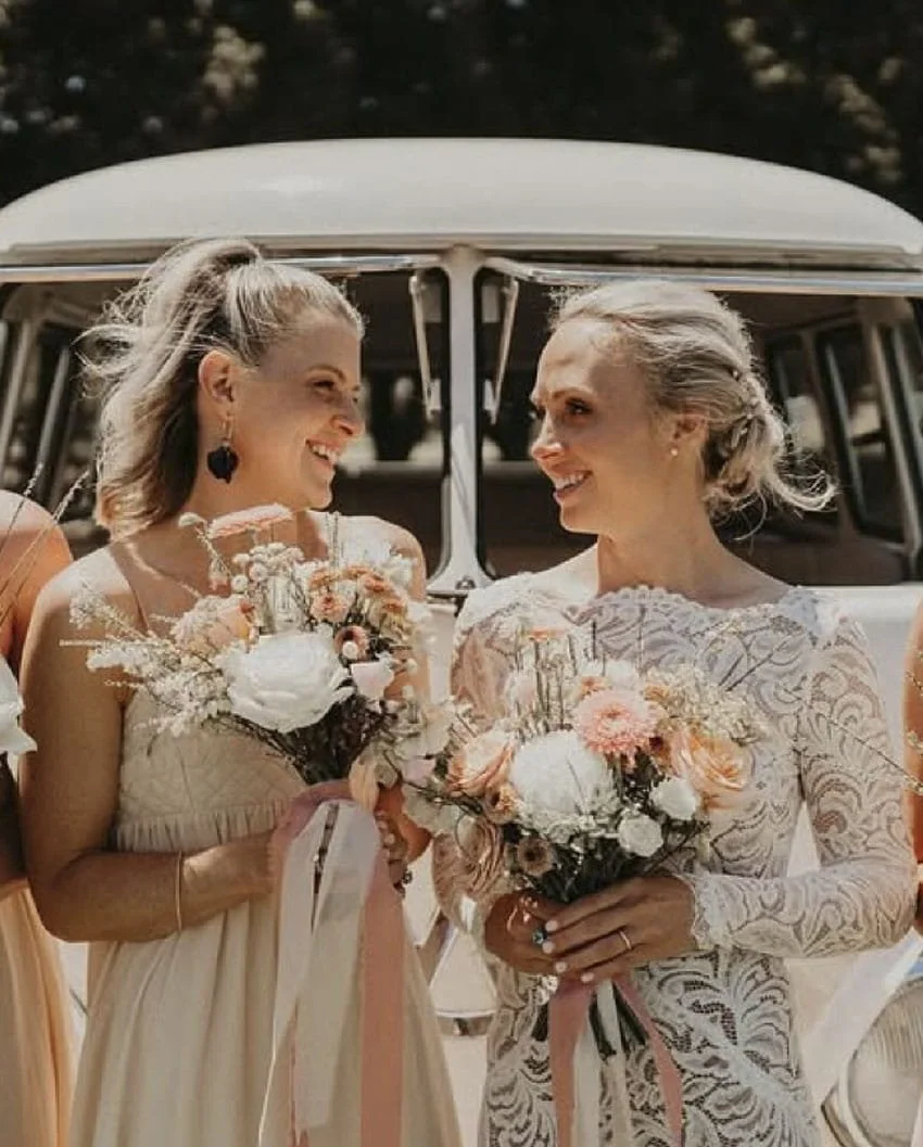 Two women in wedding dresses smiling and holding bouquets of flowers, standing in front of a vintage car.