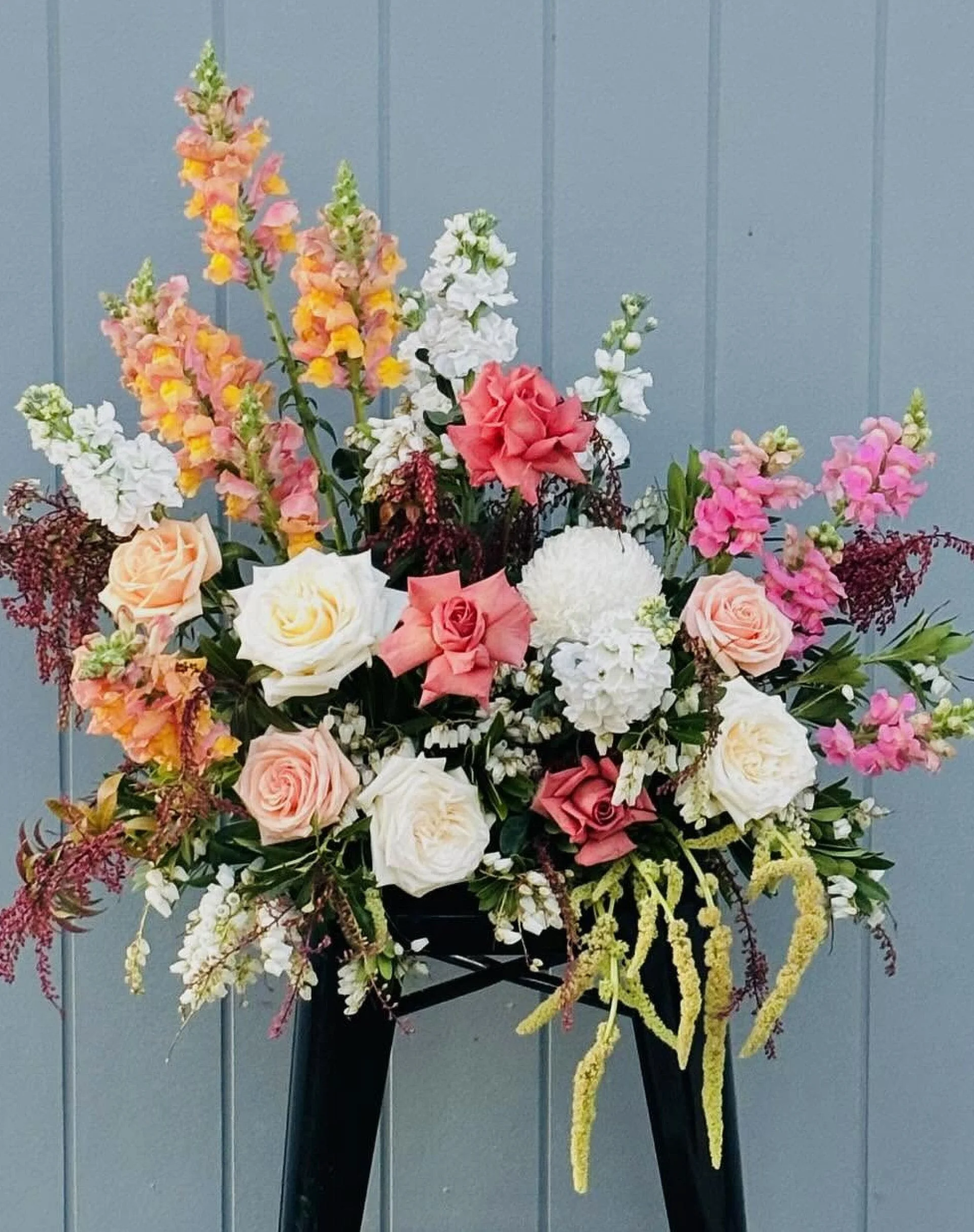 A floral arrangement with pink, white, and peach roses, snapdragons, and other flowers against a blue wall background.