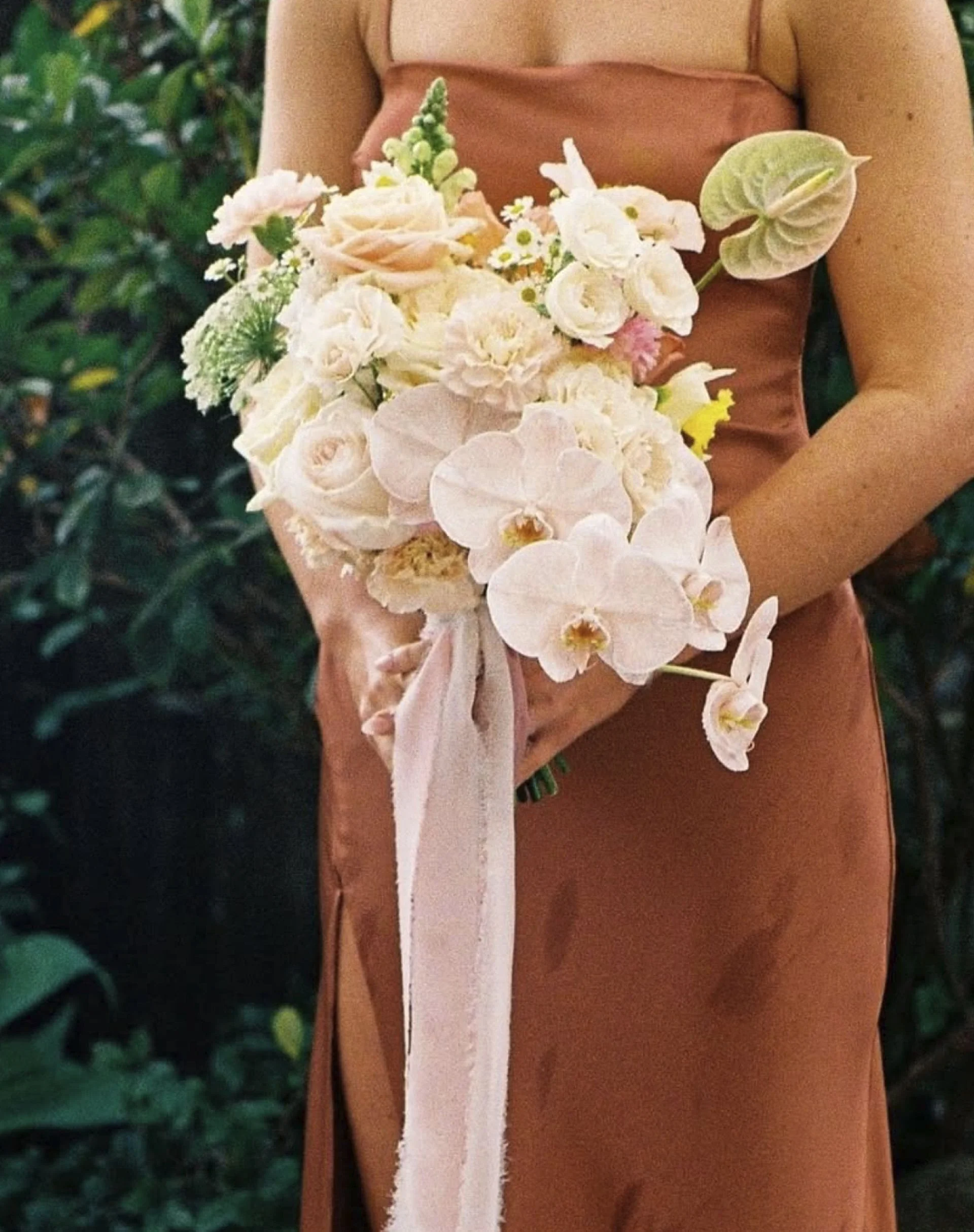 A person in a sleeveless dress holding a bouquet of white and pale pink flowers, including roses, orchids, and greenery, against a background of green foliage.