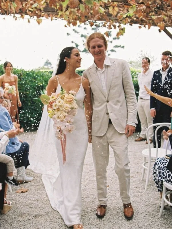 A couple in wedding attire walking down the aisle at an outdoor wedding reception, with guests and greenery in the background.