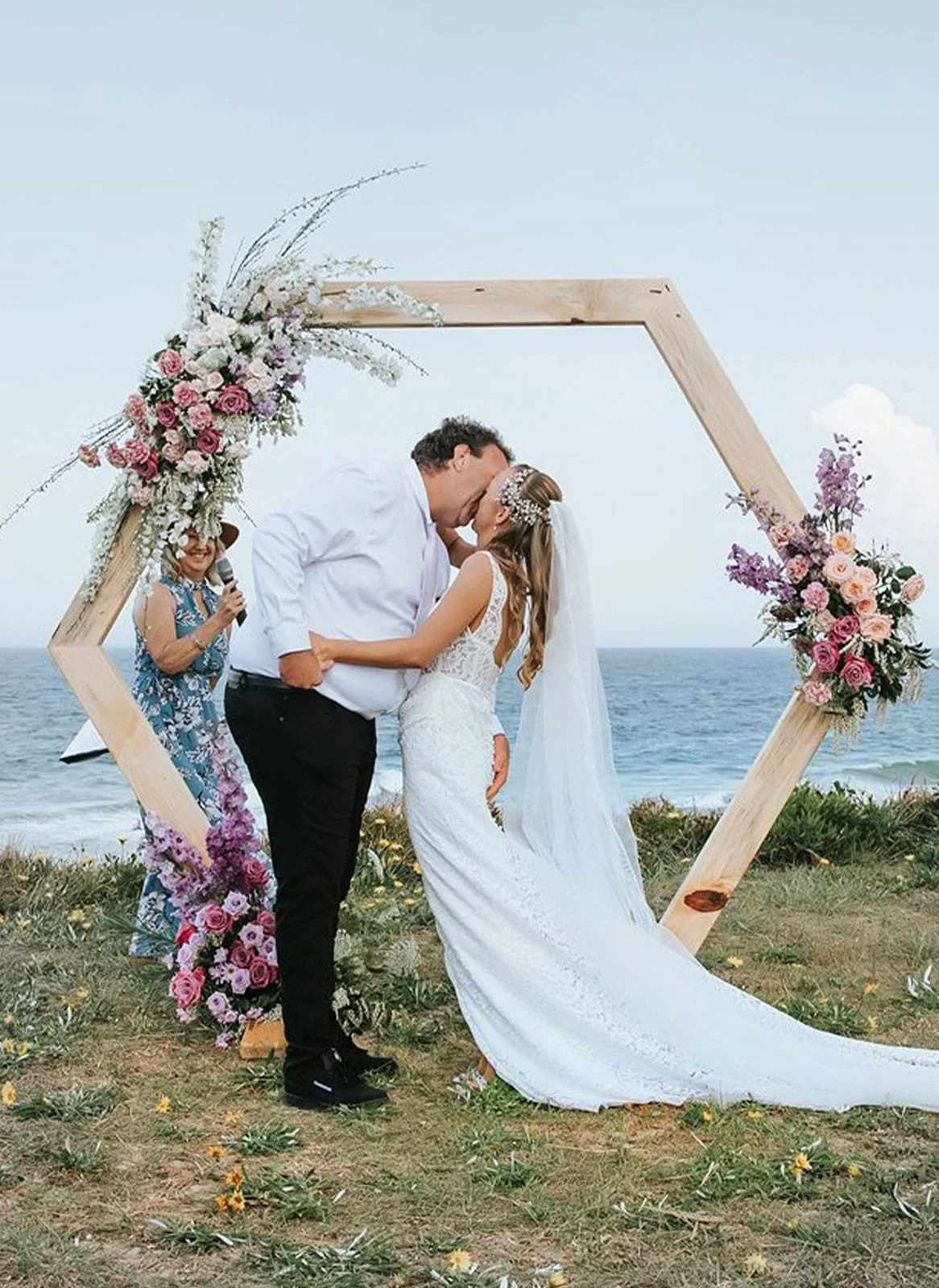 A couple getting married on a beach, kissing under a hexagonal wooden arch decorated with pink and purple flowers. An officiant stands nearby, holding a microphone.
