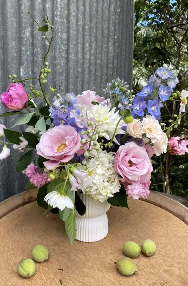 A floral arrangement with pink roses, white hydrangeas, blue delphiniums, and other small flowers in a white ceramic vase on a wooden table with green apples around it.