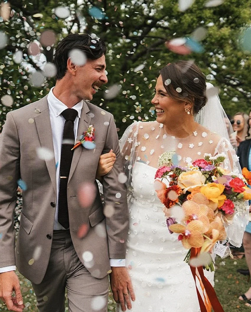 A bride and groom smiling and walking through falling confetti outdoors, surrounded by trees, with guests in the background.