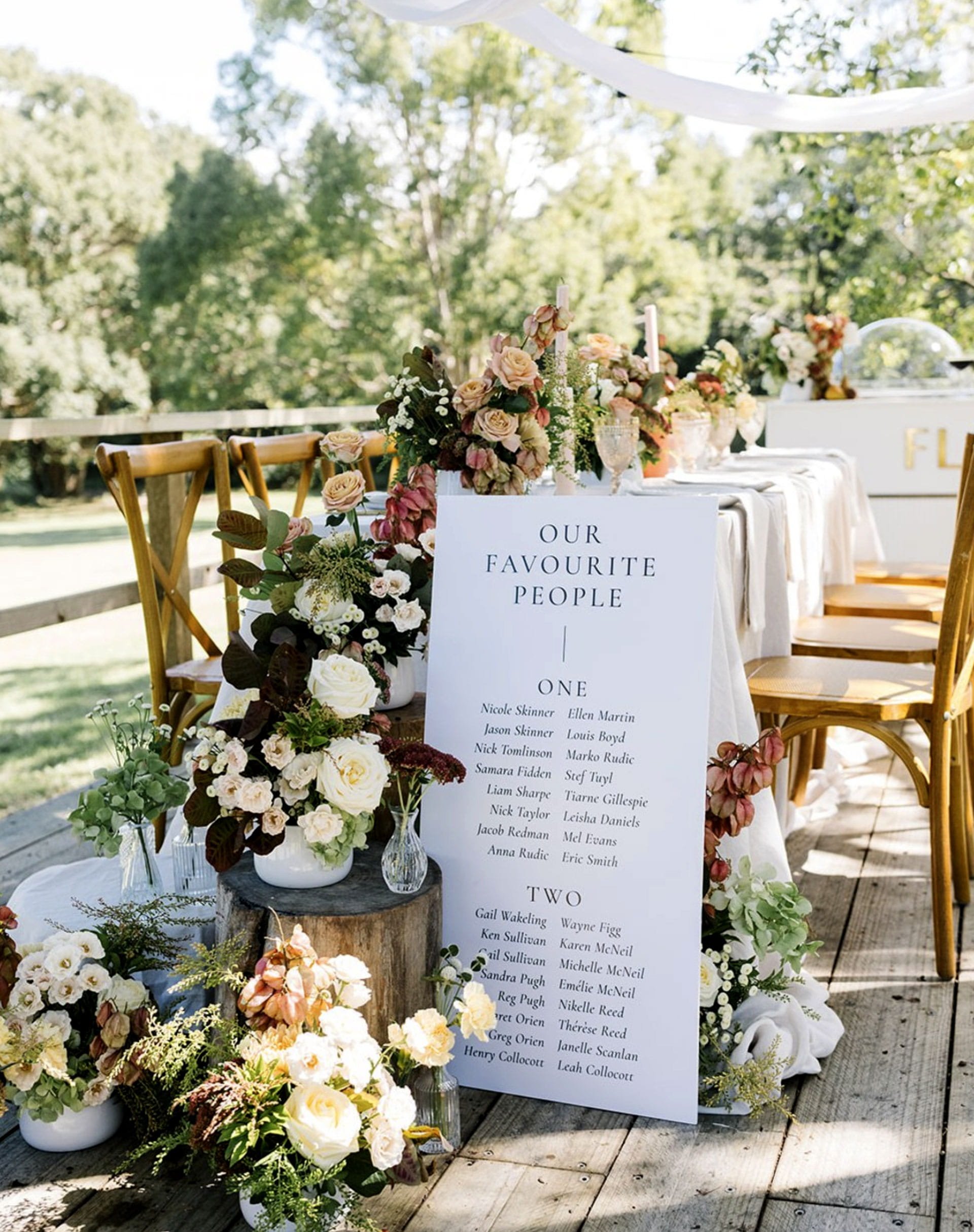 A decorated outdoor table with flowers and a seating chart for a wedding or event. The table has a sign that says 'OUR FAVOURITE PEOPLE' with a numbered list of names below, and there are various floral arrangements in vases on the table and around i