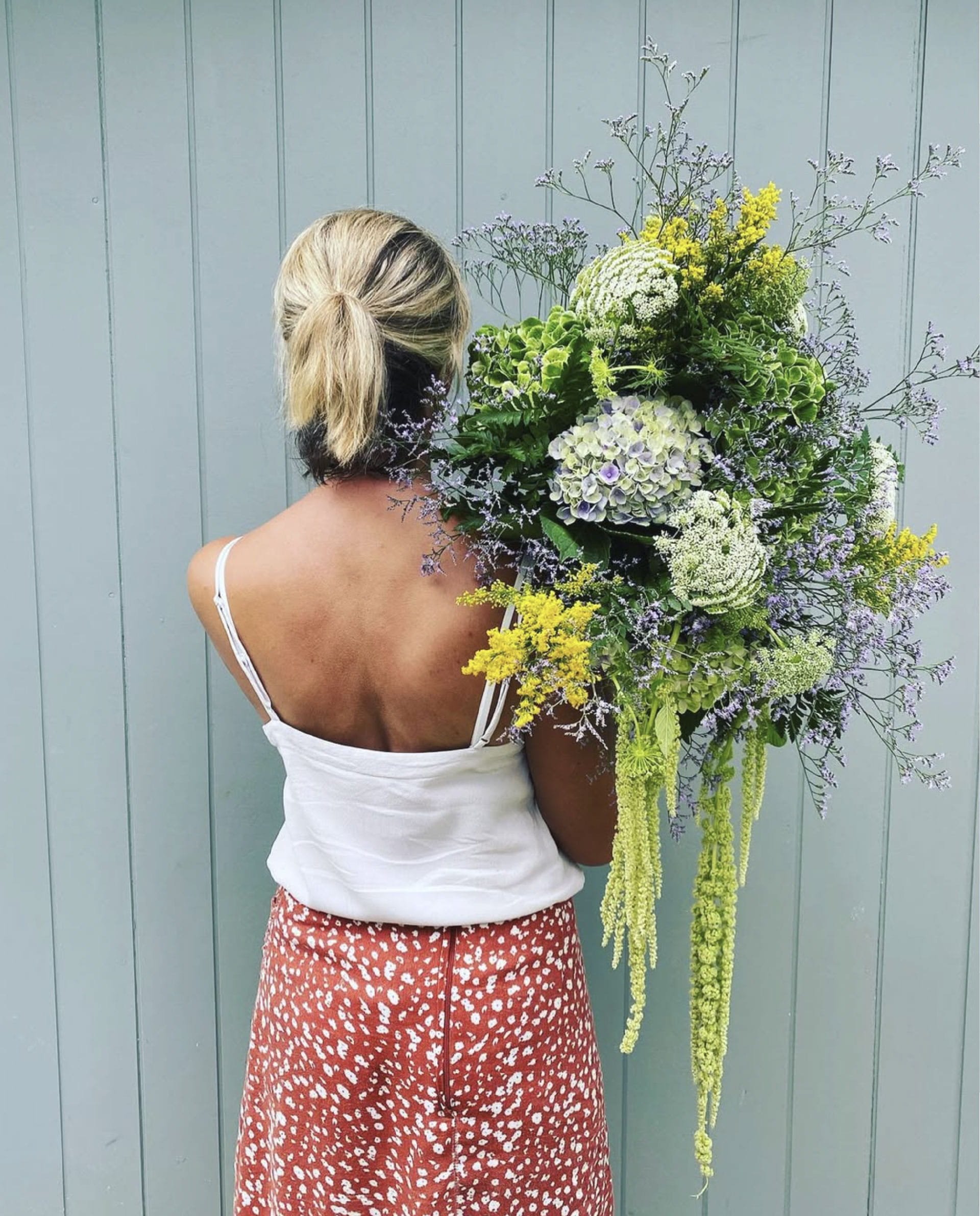 A woman with blonde hair tied in a ponytail, wearing a white top with spaghetti straps and a red skirt with white spots, is holding a large bouquet of assorted flowers while standing against a light blue wooden panel wall.