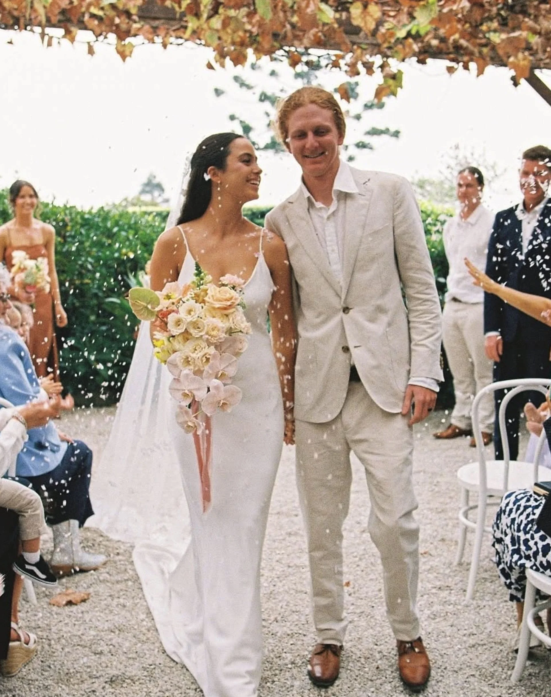 A bride and groom smile as confetti falls during their wedding celebration outdoors. The bride wears a white dress and holds a large bouquet of flowers. The groom is dressed in a light-colored suit and brown shoes. Guests are seated on either side, with some standing and smiling in the background, and some children sitting on chairs. The setting is decorated with greenery and a wooden arch.