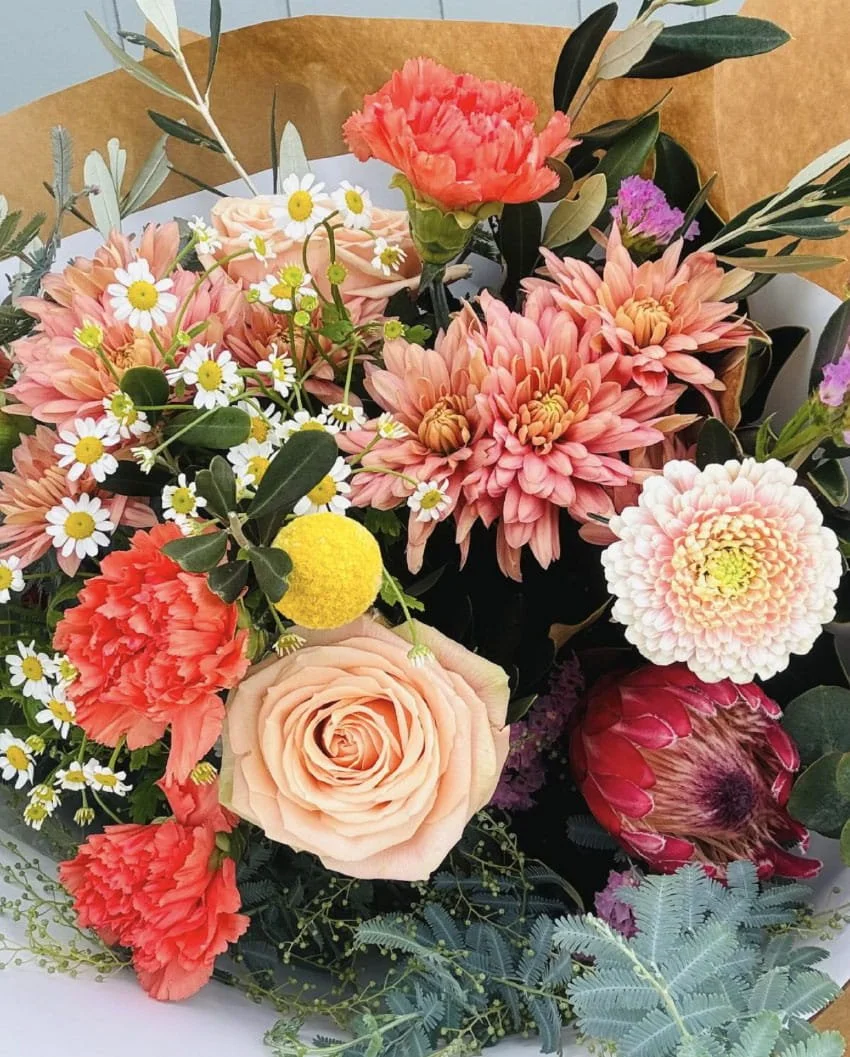 A colorful bouquet of various flowers including pink carnations, white daisies with yellow centers, peach roses, peach dahlias, and a red protea, with greenery and brown wrapping paper in the background.