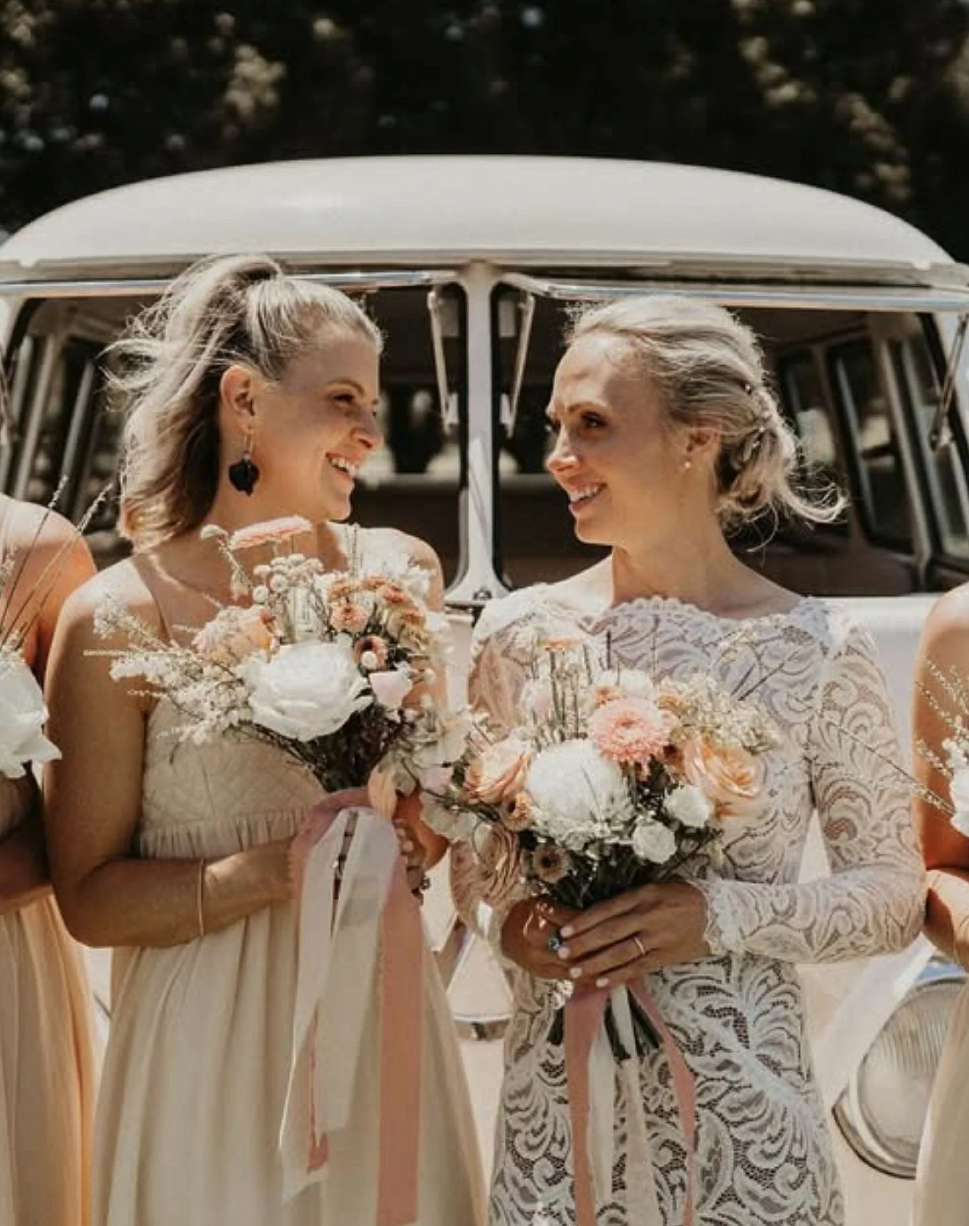 Two brides with blonde hair in wedding dresses holding bouquets, smiling at each other inside a vehicle.