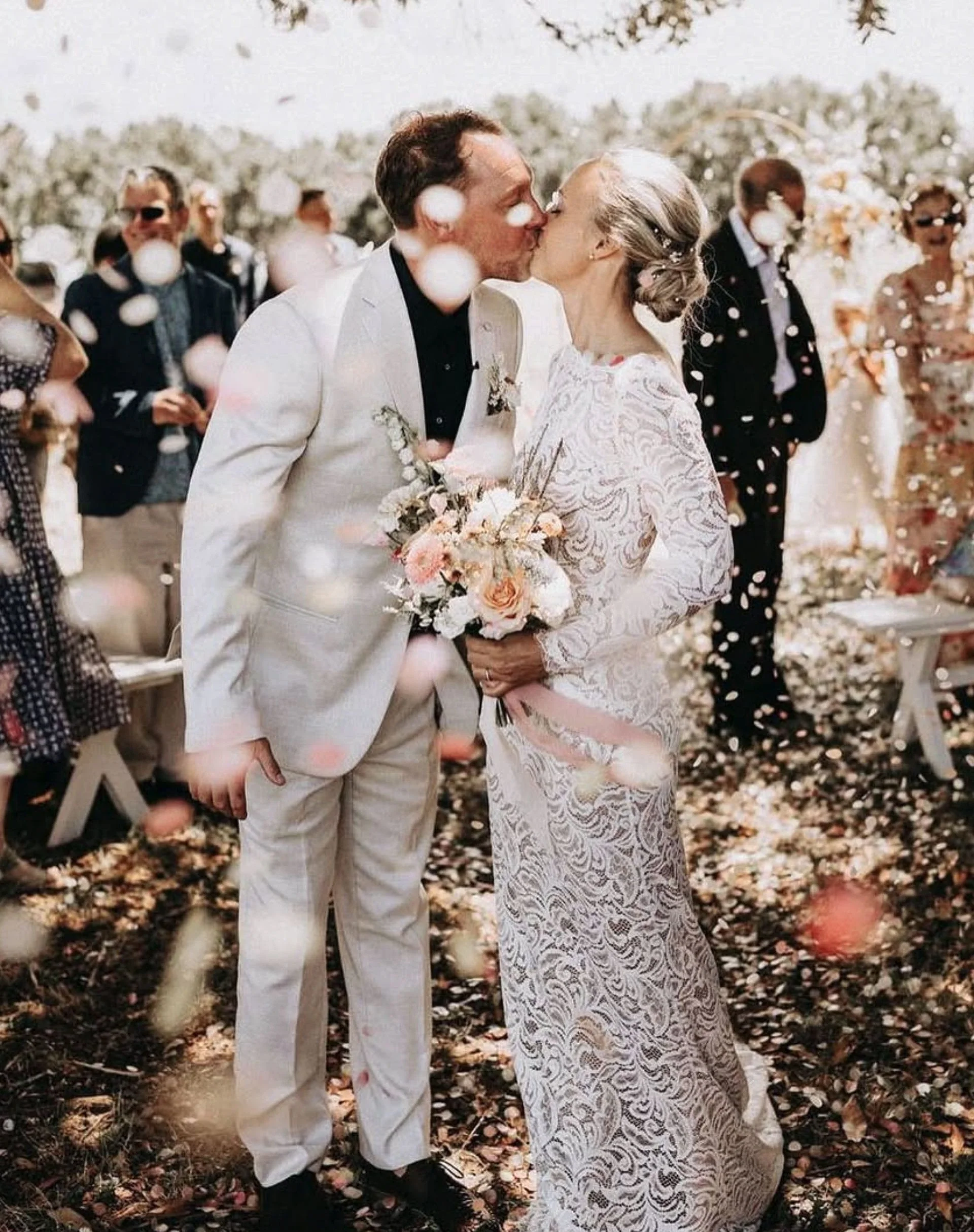A woman and a man kiss during a wedding ceremony outdoors surrounded by guests and fallen autumn leaves.