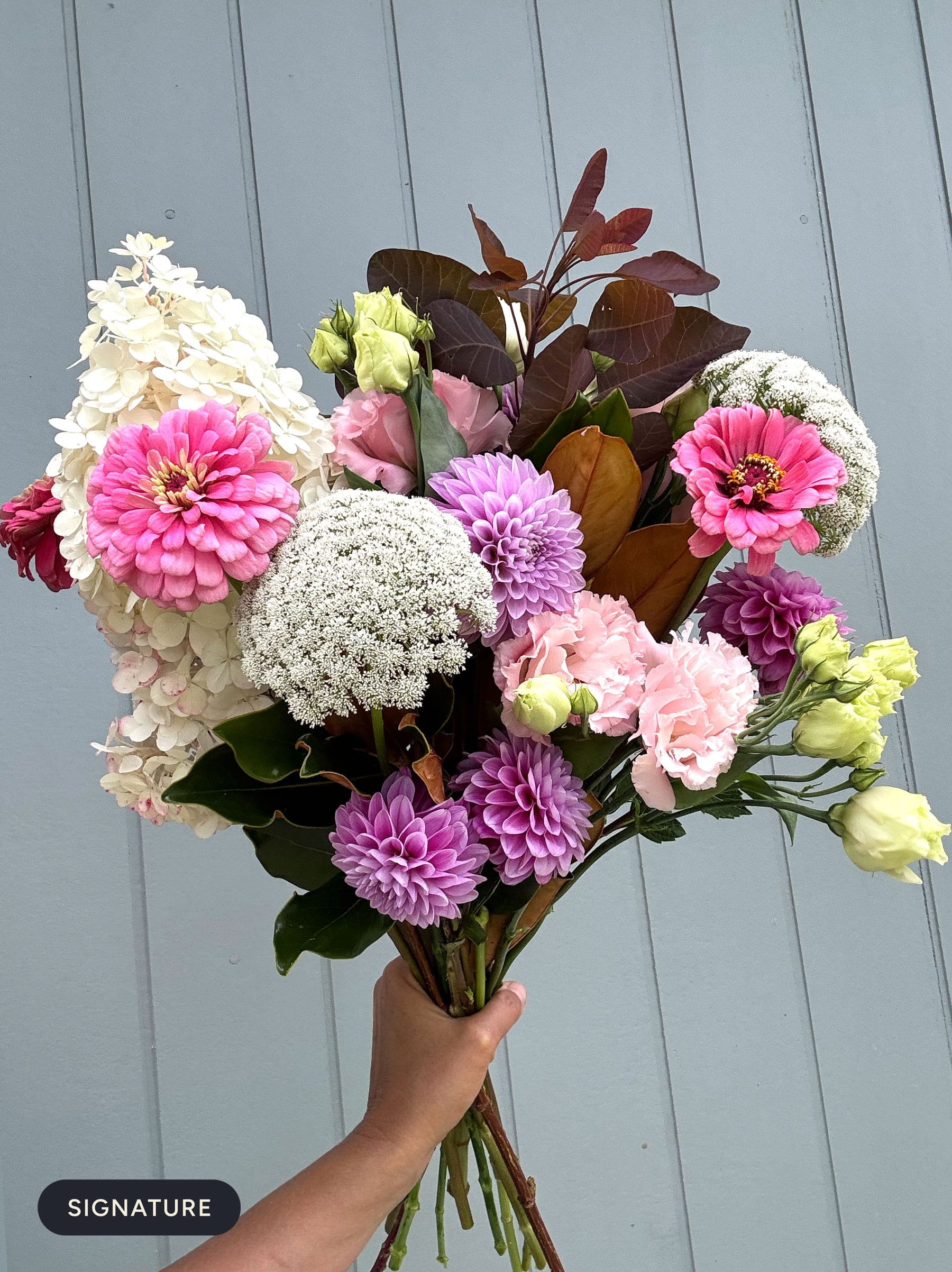 A hand holding a bouquet of various colorful flowers against a light gray wooden background.