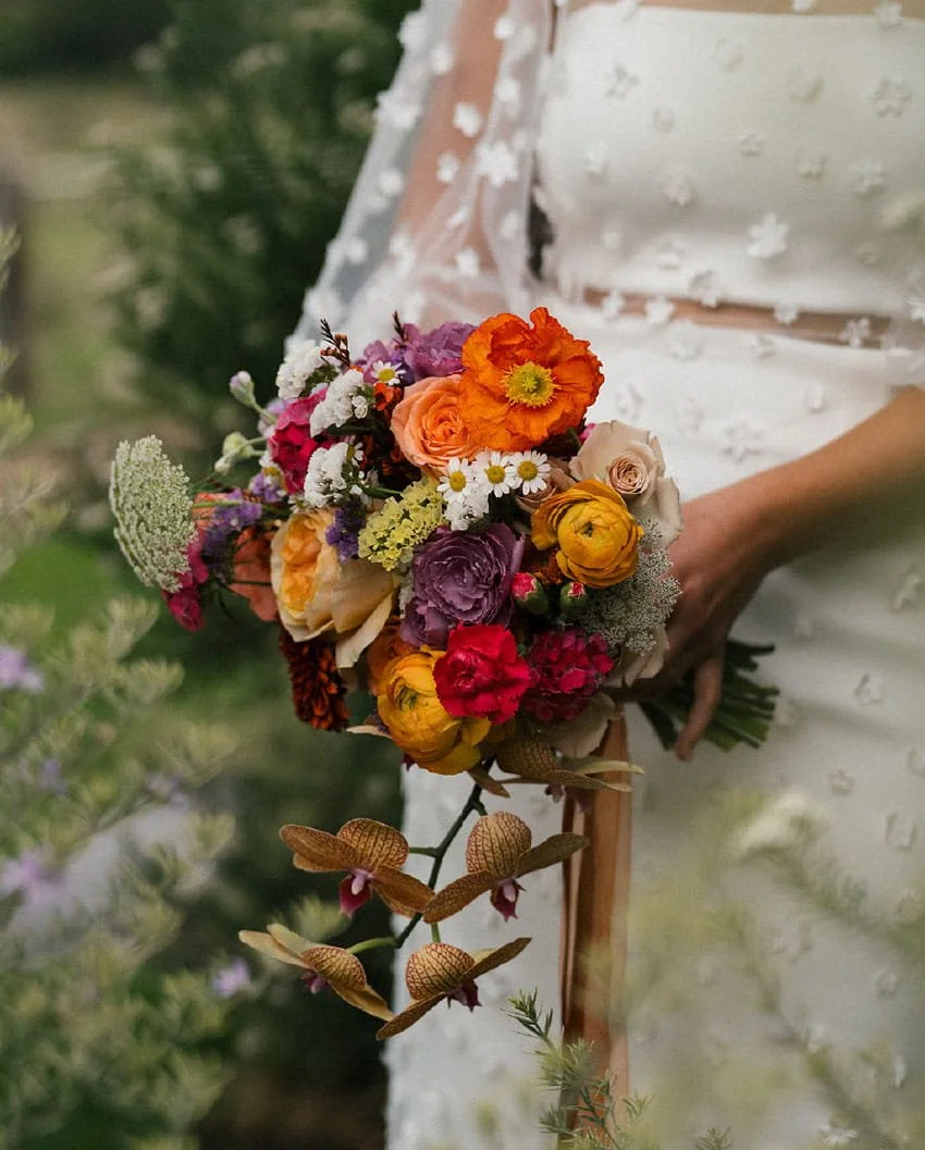 Close-up of a woman holding a colorful bouquet of flowers, including orange, purple, pink, and white blossoms, with a white dress featuring floral embroidery.