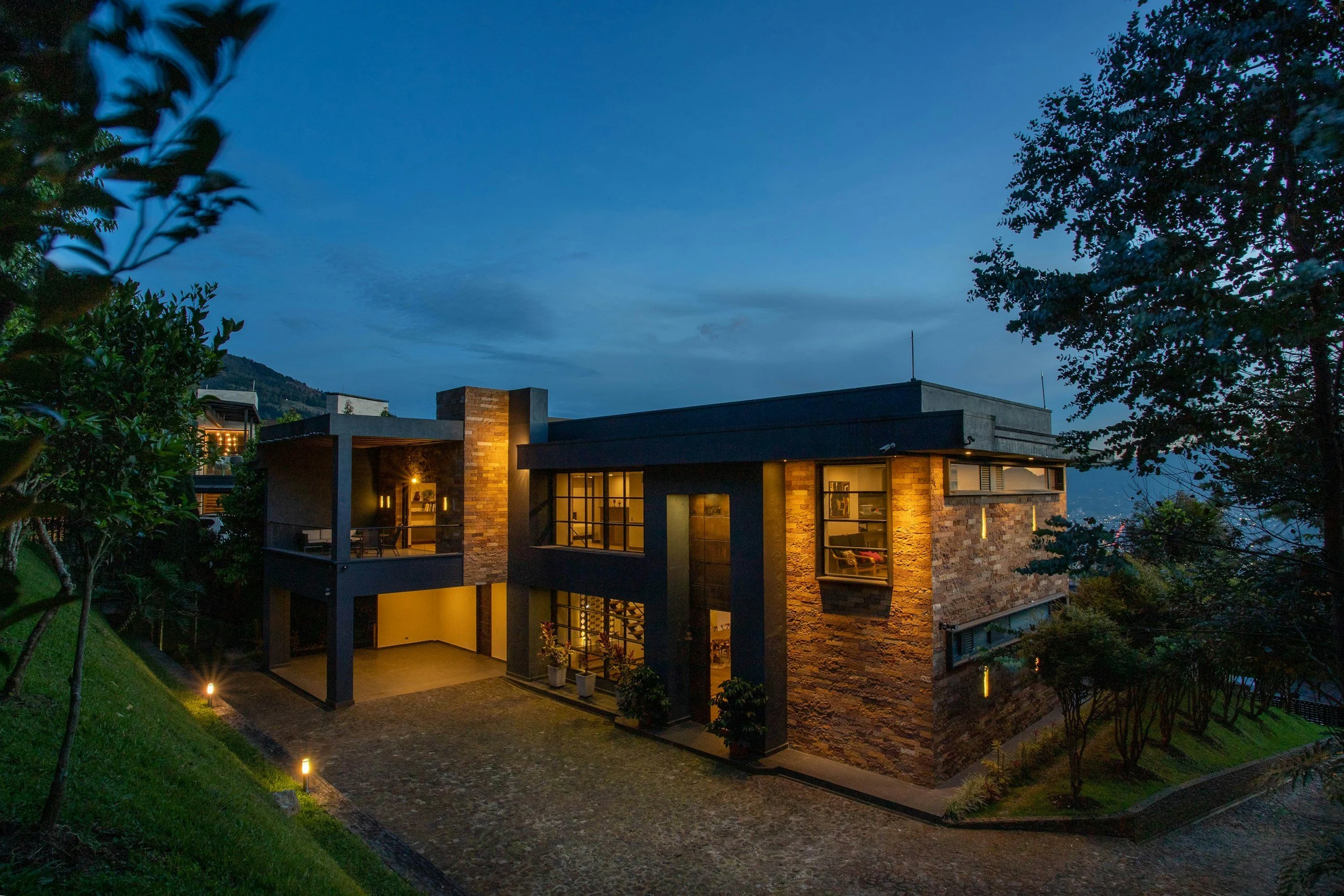 Modern house with large glass windows, brick and dark gray exterior, illuminated from within, situated on a landscaped hillside with trees and pathway at dusk.